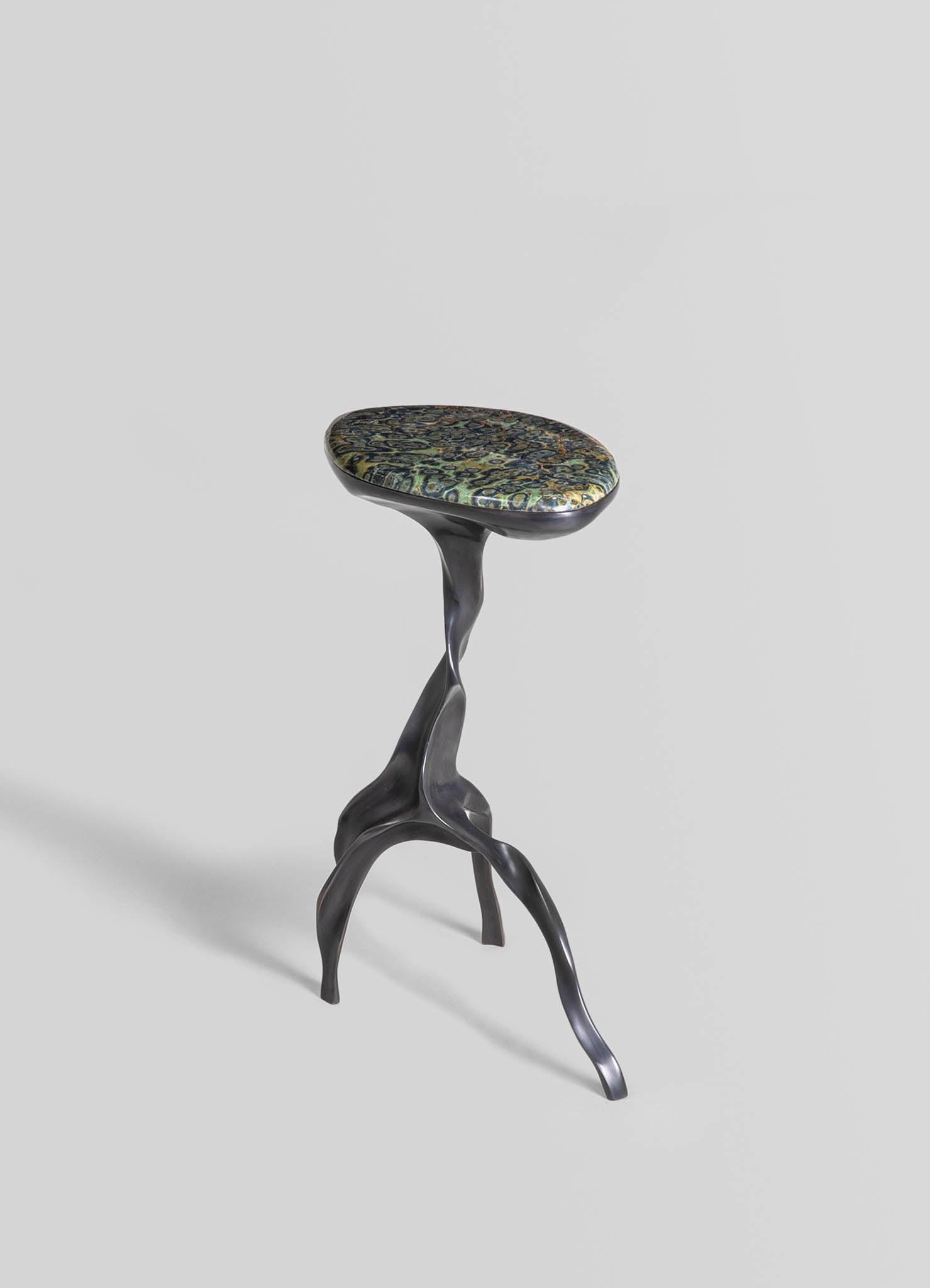 A sculptural side table with a dark, wavy metal base resembling natural forms and an oval top featuring a colorful, intricate pattern in shades of green, blue, and gold. The design combines organic shapes with decorative motifs.