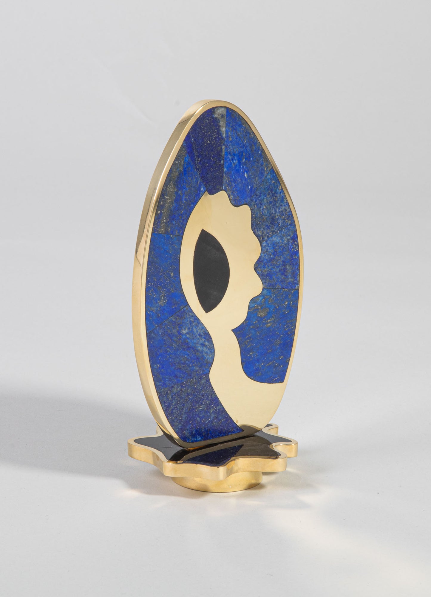 A decorative table sculpture featuring a semi-abstract face in cream-colored material, set against a bold blue background with gold accents and black details. The piece has a contemporary style with smooth, rounded shapes and contrasting colors.
