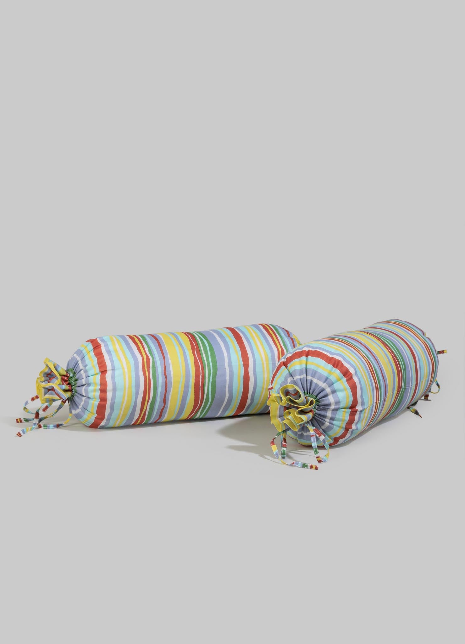 Two fabric bolster cushions with multicolored vertical stripes and fabric ties, made of lightweight cotton or similar material, placed on a plain surface.