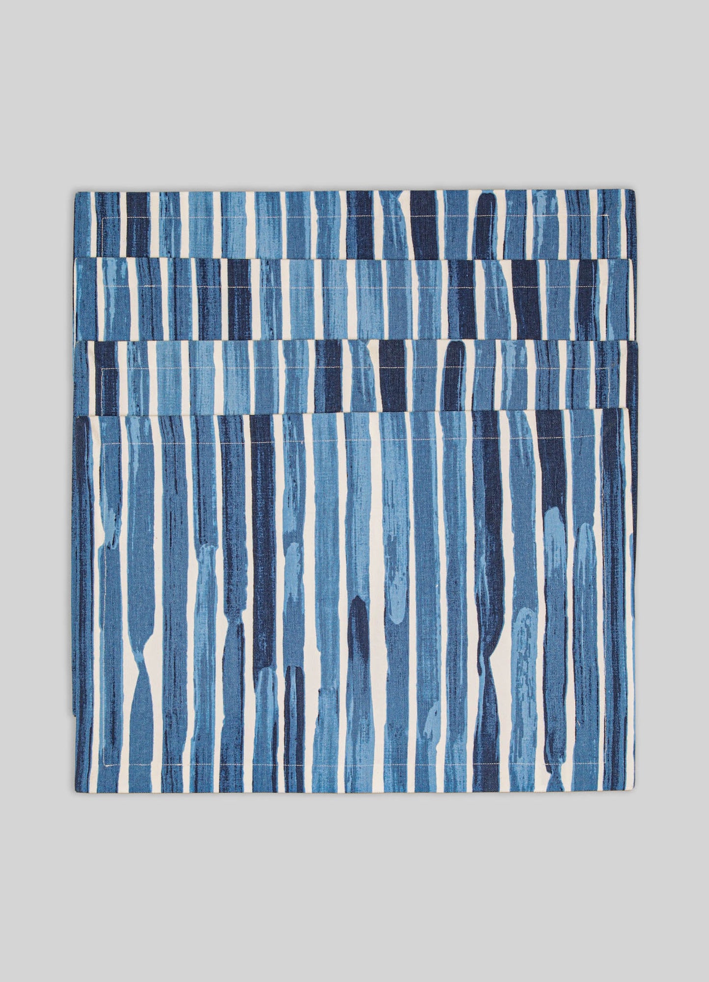 A fabric napkin featuring a pattern of vertical brushstroke-like stripes in varying shades of blue, set against a neutral gray background. The textile appears to be made of cotton or linen and has a contemporary, casual style with an abstract motif.