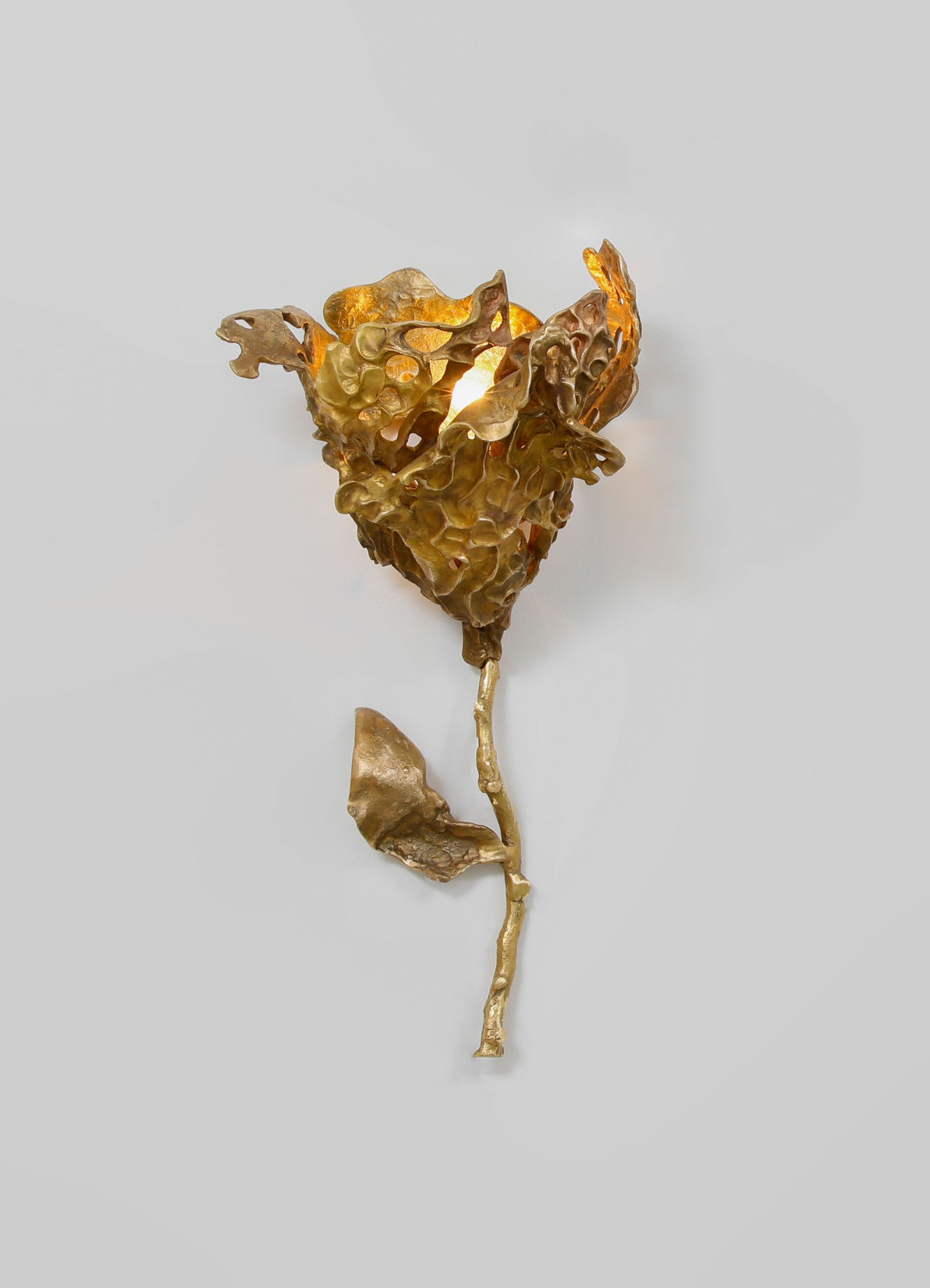 A metallic gold wall sconce with an organic, textured design resembling a ruffled flower or leaf, with a light bulb illuminating from within. The fixture has a sculptural, contemporary style with intricate, irregular details.