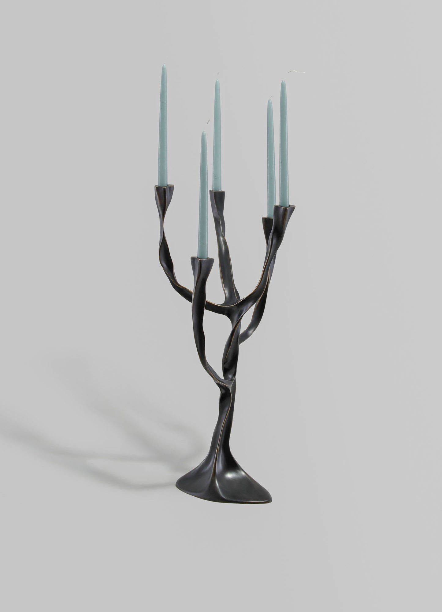 A sculptural candle holder with a black, twisted metal base and five light blue tapered candles, designed with an organic, flowing form on a plain light gray background.