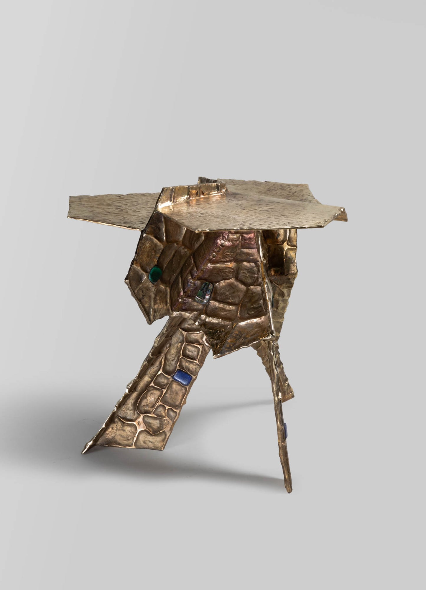 A sculptural side table crafted from hammered and textured metal with a distressed gold finish, featuring irregular, geometric shapes and embedded colored glass stones, set against a plain background.