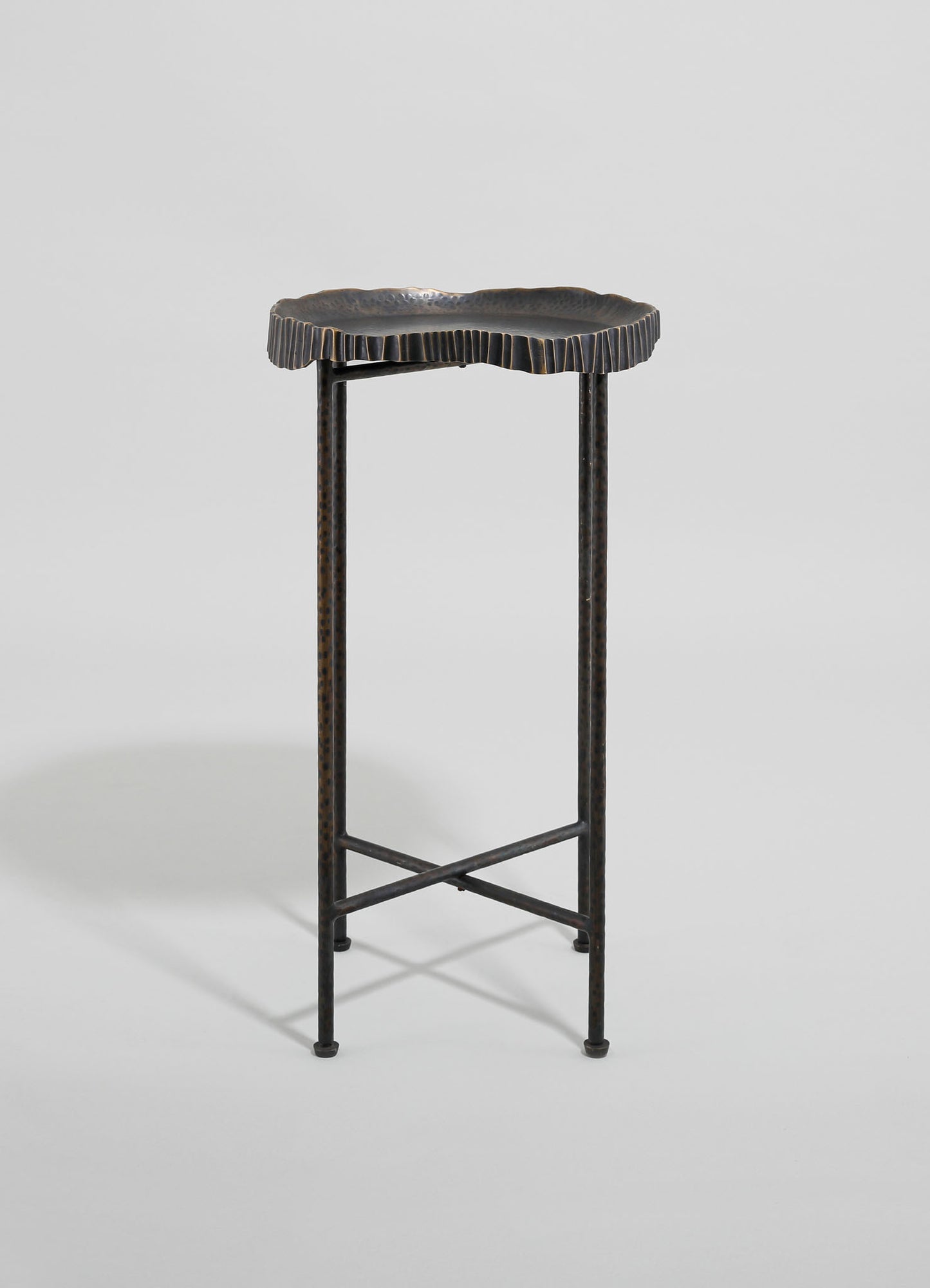 A metal side table with a textured, irregular-shaped top and slender, perforated legs connected by crossbars. The finish appears dark and matte, emphasizing a handcrafted, rustic style.