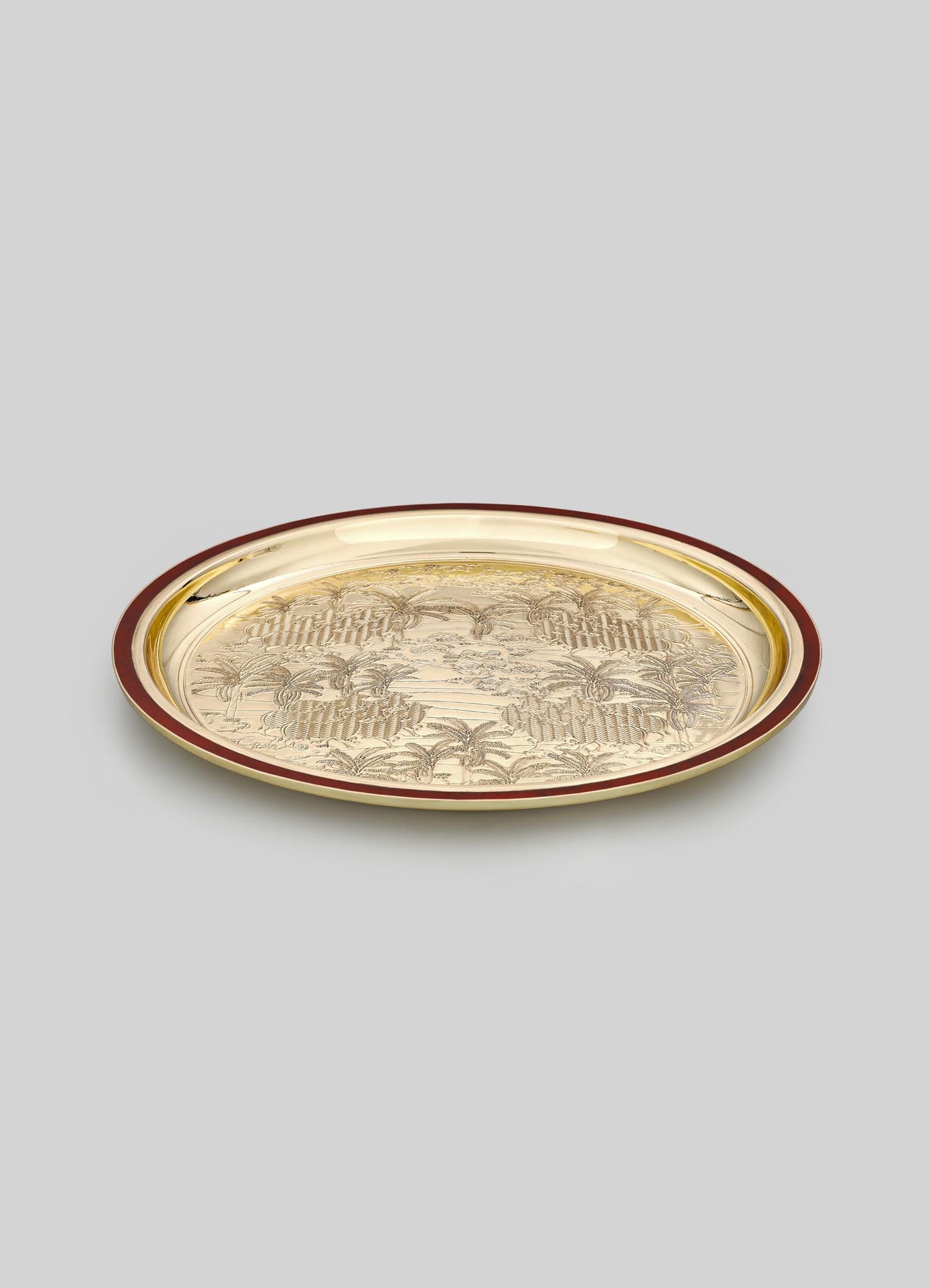 A round metal tray with an intricate engraved landscape motif featuring palm trees and a decorative border, finished in gold with a contrasting dark red rim.