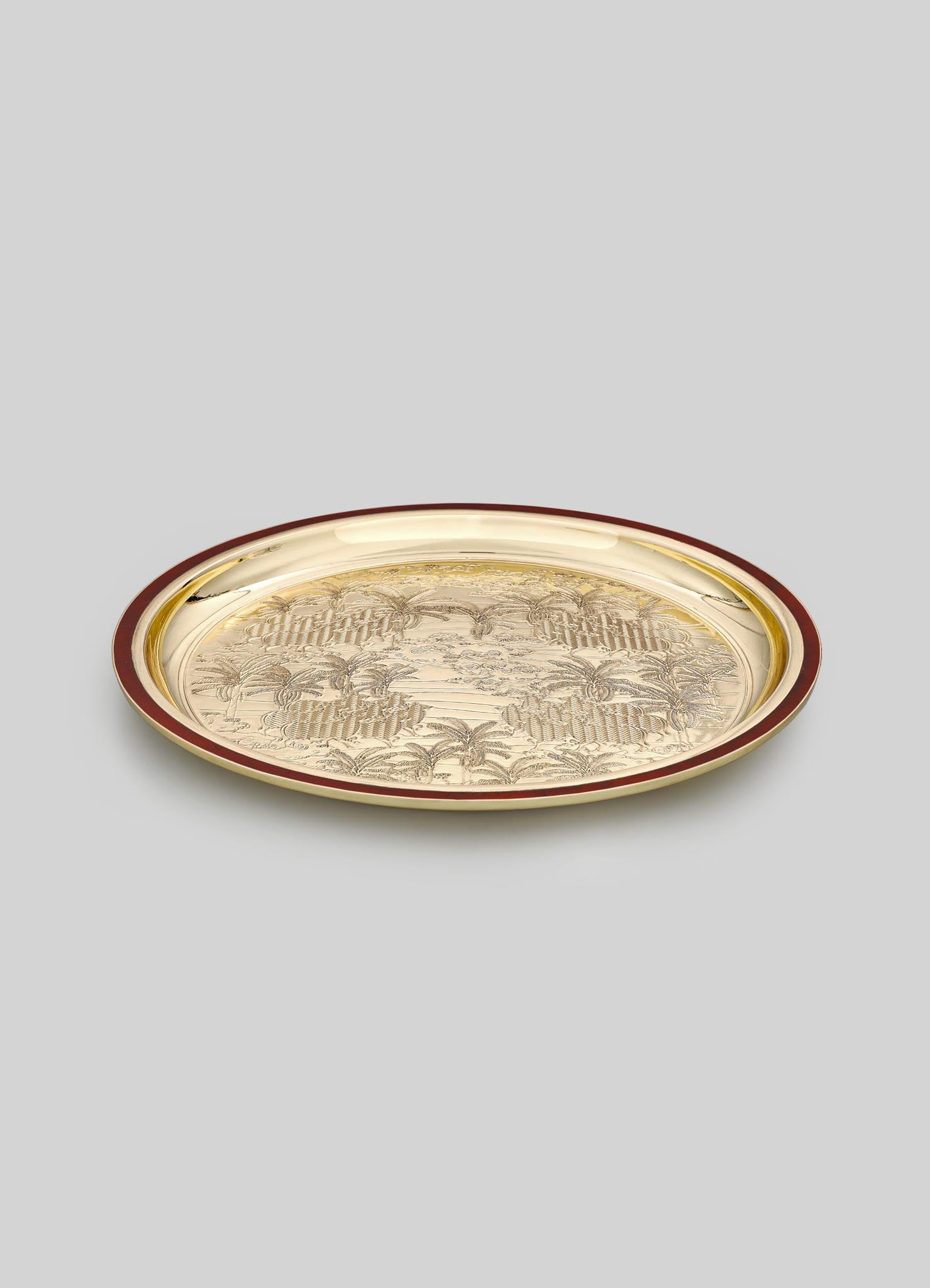 A round metal tray with an intricate engraved landscape motif featuring palm trees and a decorative border, finished in gold with a contrasting dark red rim.