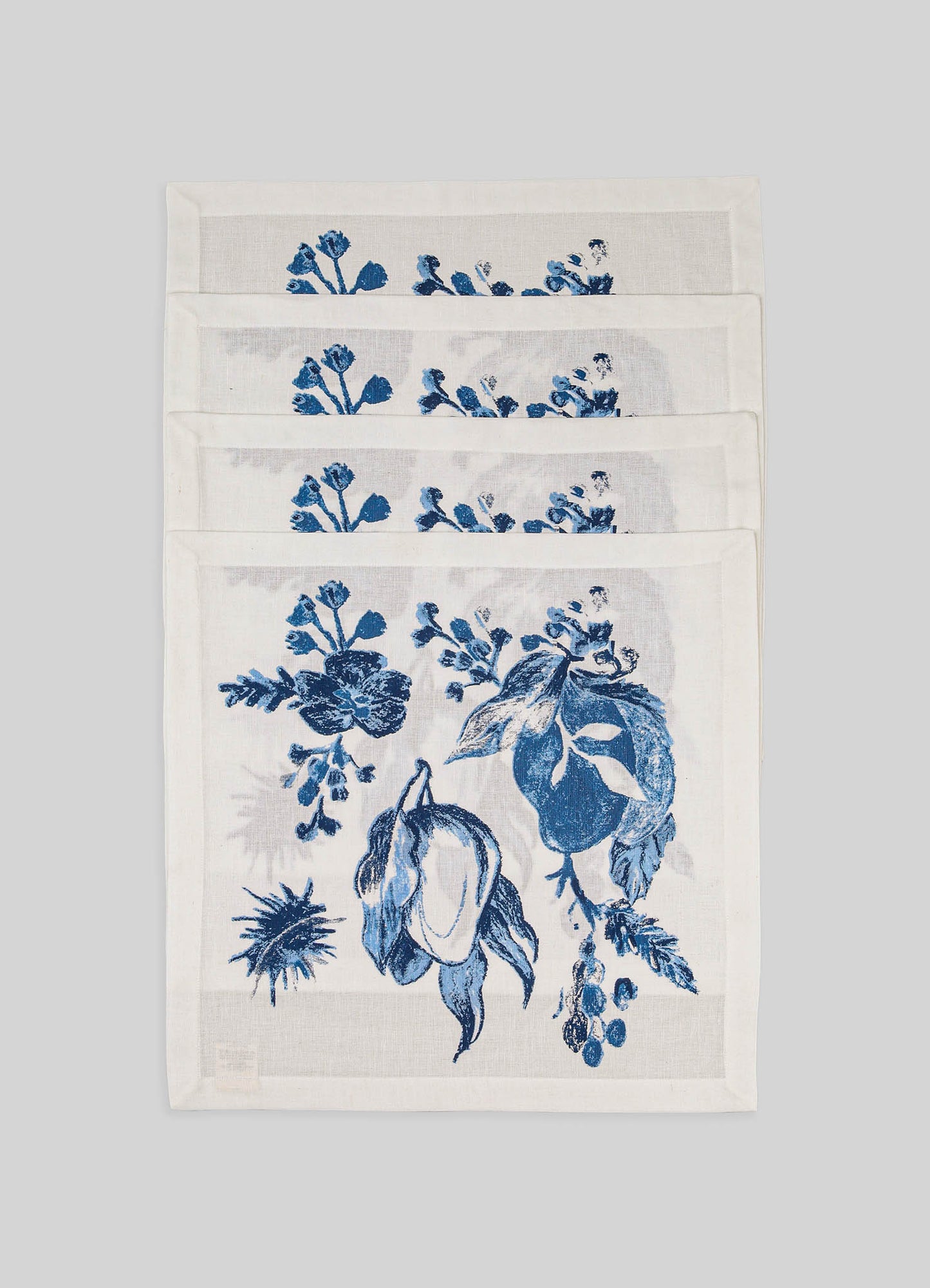 Stacked linen napkins with blue floral and fruit motifs on a white background, featuring a hand-printed design and clean, simple borders.