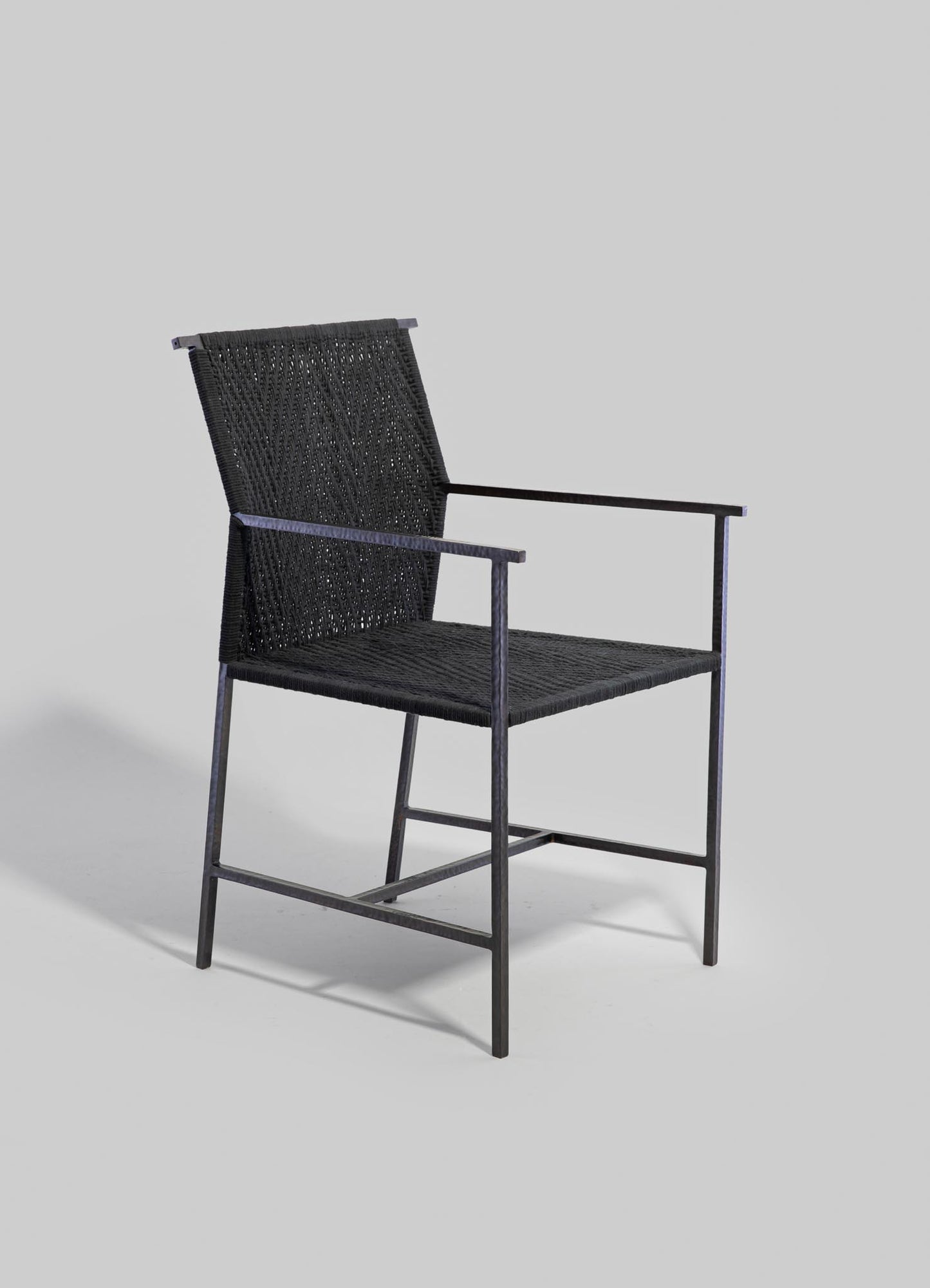 A black woven rattan or wicker chair with a metal frame, featuring a tall backrest and armrests, in a contemporary style. The materials include textile or synthetic fibers for the seat and back, and metal for the structure. The design is minimalist with a sleek, modern aesthetic.