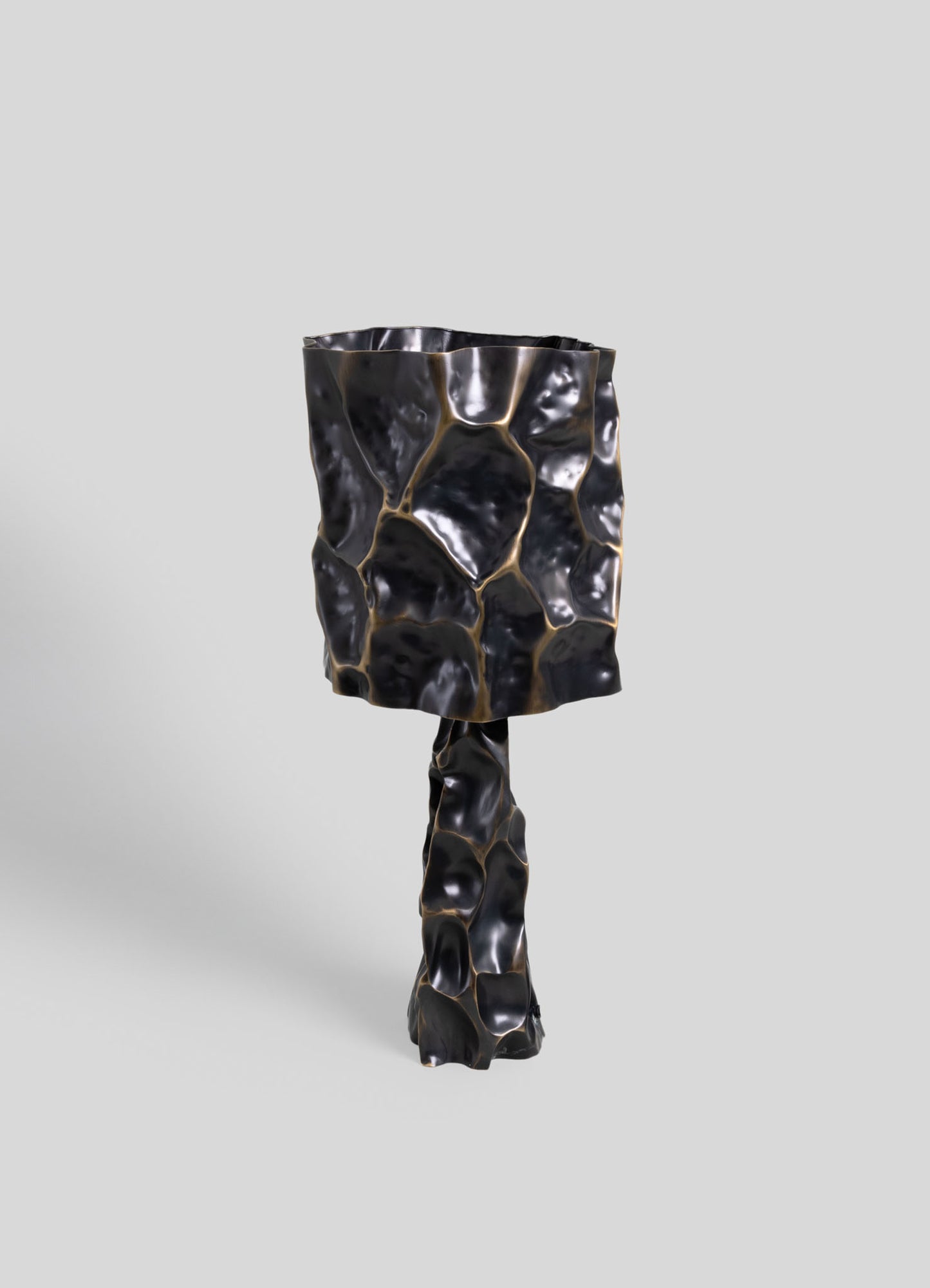 A sculptural table lamp with a textured, organic pattern resembling cracked or fractured stone, finished in dark metallic with gold accents, set on a matching base. The design has a contemporary and handcrafted aesthetic with a bold, sculptural presence.