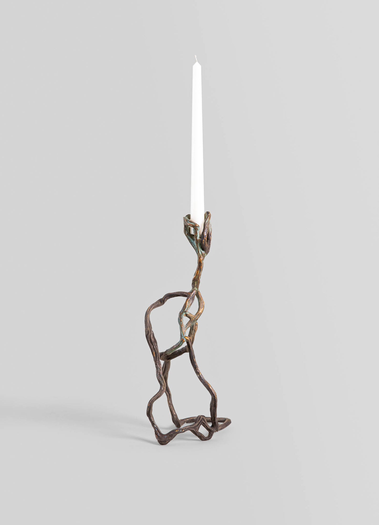 A decorative candle holder made of twisted metal with a rustic, bronze finish, holding a tall, white tapered candle. The design features organic, irregular shapes with an artistic, handcrafted appearance.