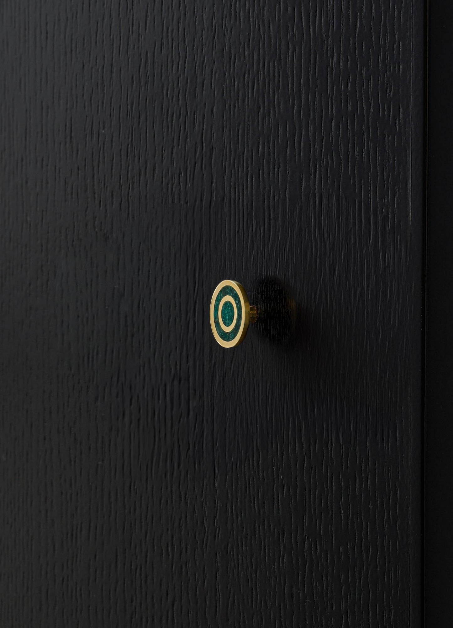 A black wooden surface featuring a decorative round knob with concentric circles in turquoise and white, framed by a slim gold border. The finish of the wood is textured with visible grain patterns.