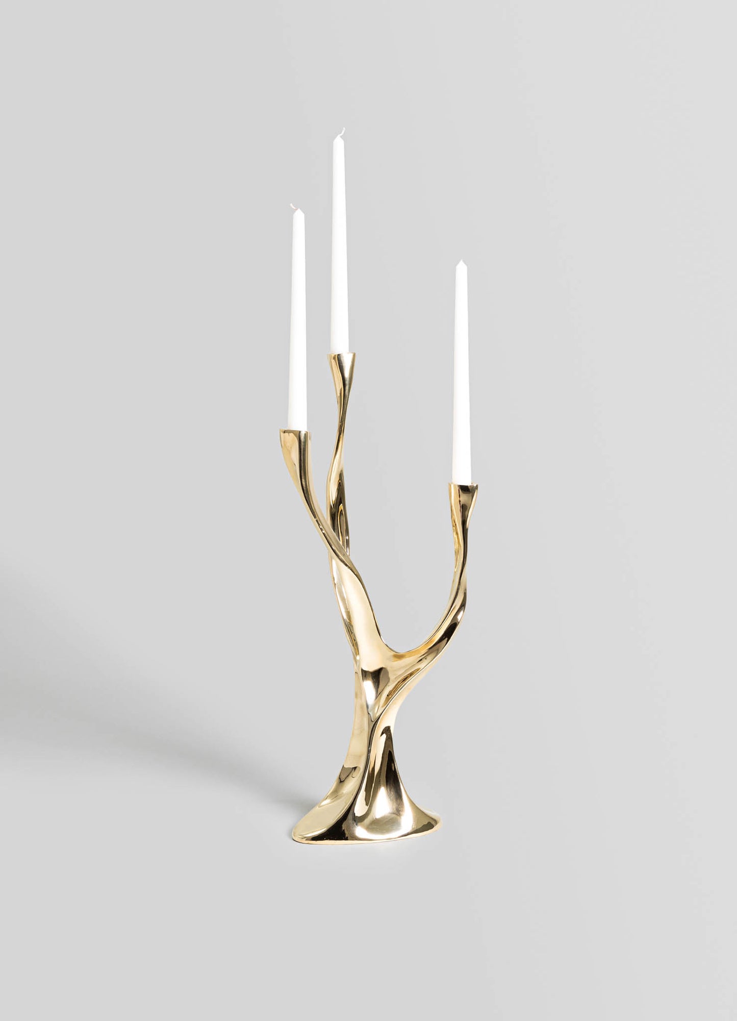 A decorative candelabrum with a sculptural, organic design in polished gold-tone metal, holding three tall white taper candles.
