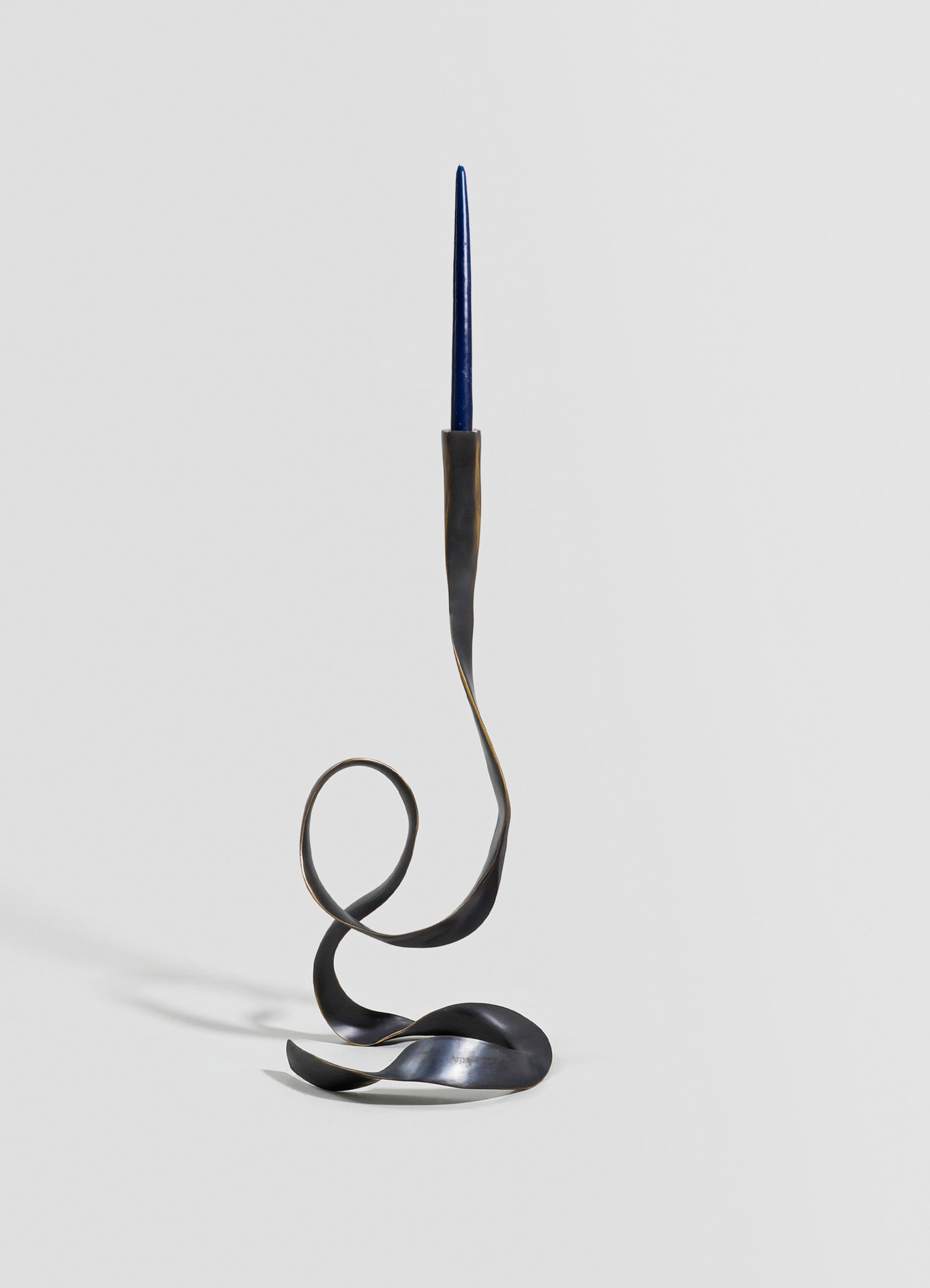 A sculptural candlestick holder made of dark metal with a twisted, flowing design, featuring a slim blue taper candle at the top. The piece has an artistic, contemporary style with organic curves and a matte finish.