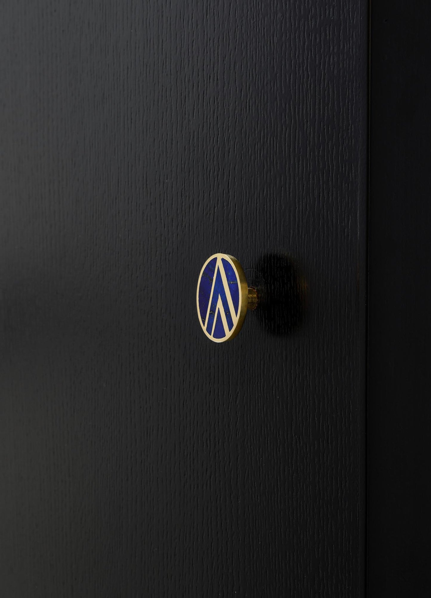 Close-up of a black textured wooden surface with a small round decorative knob featuring a blue and gold geometric pattern. The style appears modern and sleek.