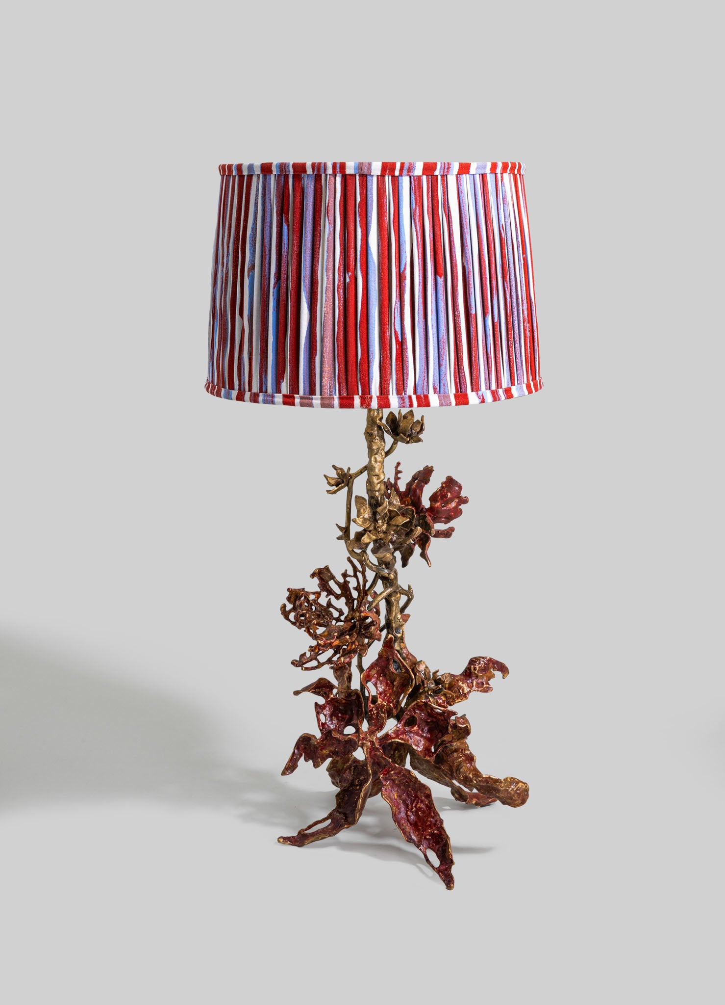 A table lamp with a sculptural base resembling coral, made of metallic material with reddish-brown and gold tones, and a fabric lampshade with vertical red, white, and purple stripes.