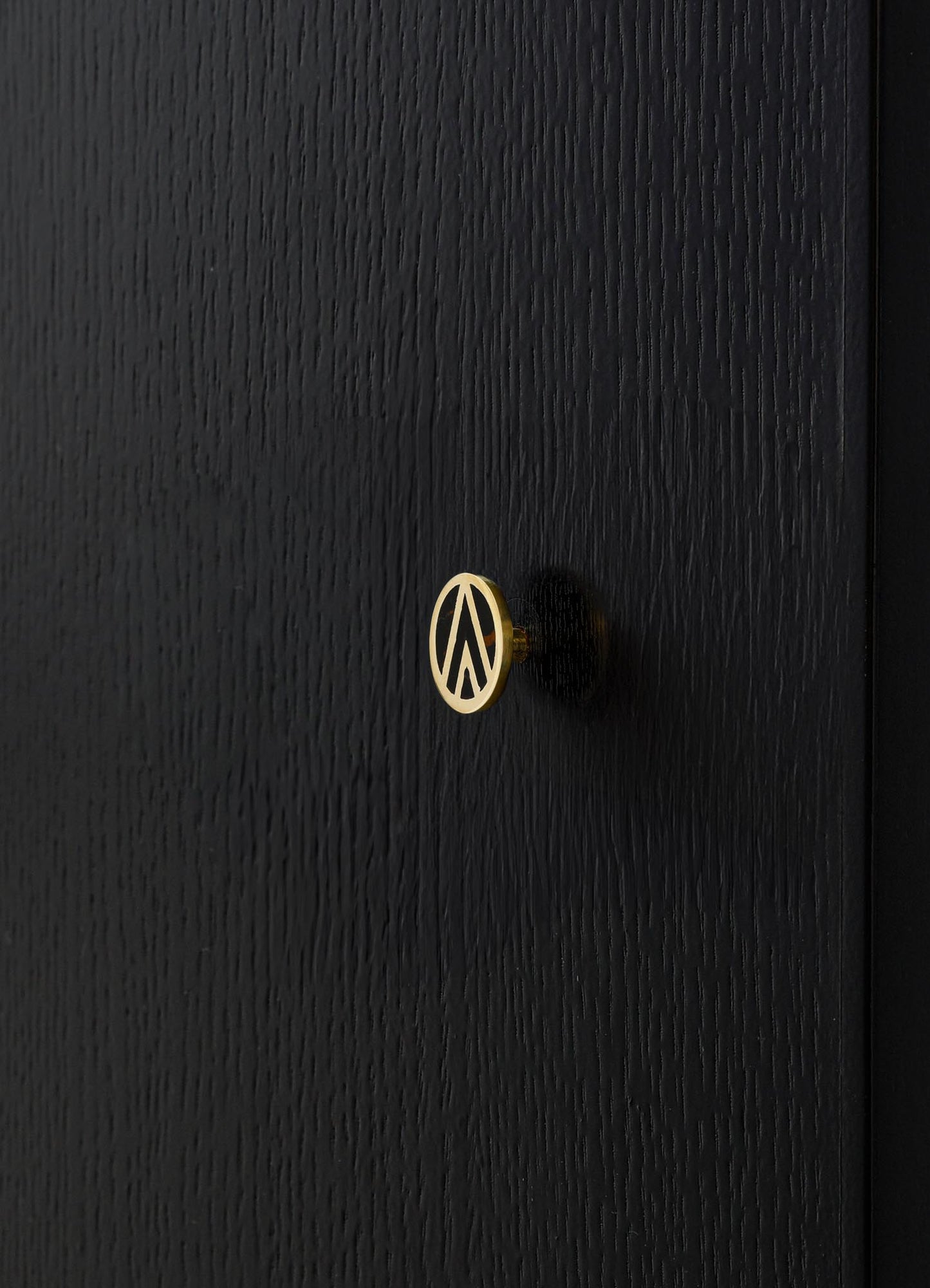 A black wooden surface featuring a small round brass knob with a geometric pattern in dark and light tones. The wood has a textured, matte finish.