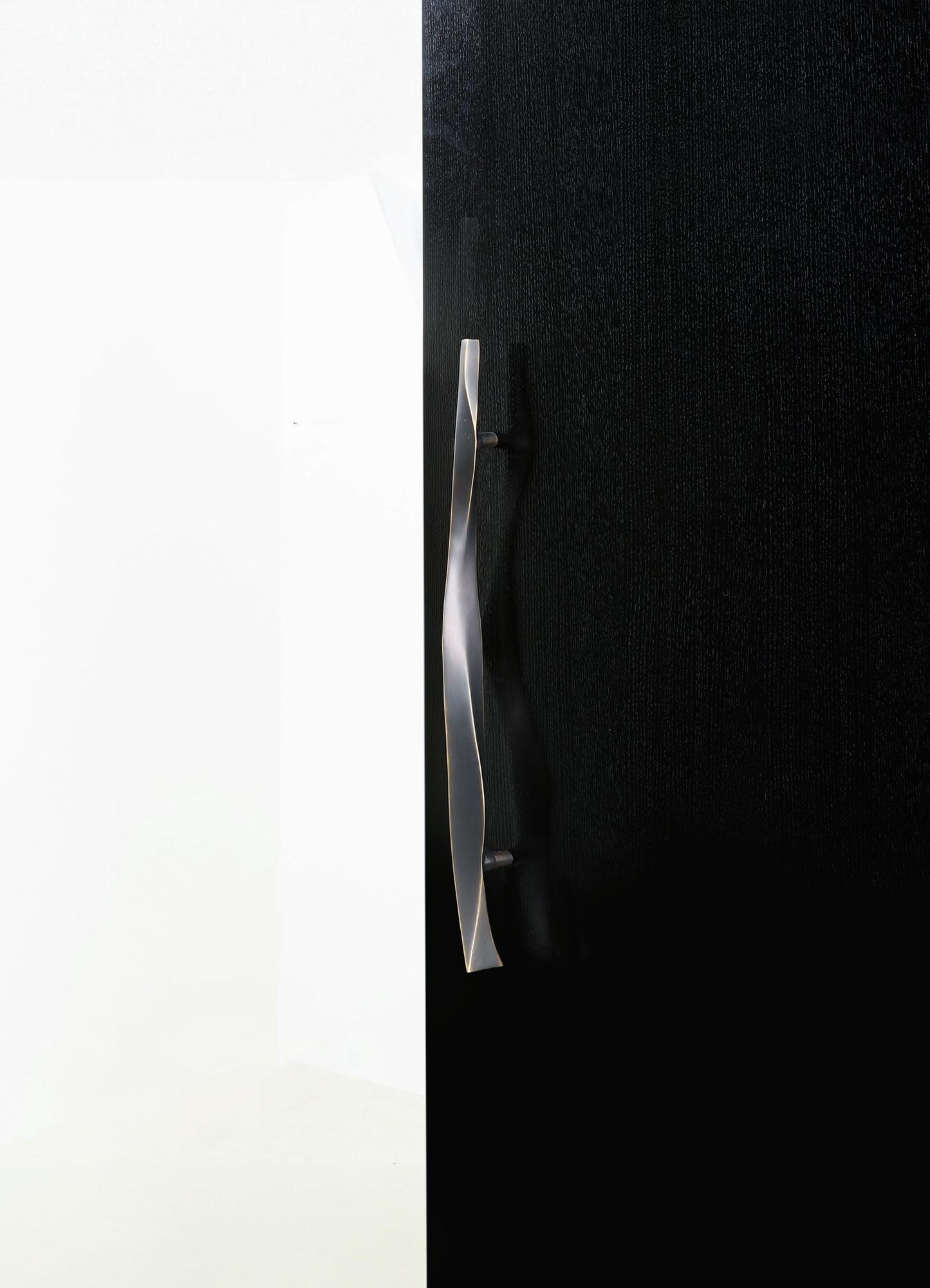 A modern door handle made of metal with a sleek, curved design, mounted on a black textured door. The handle has a polished, metallic finish and is positioned vertically.