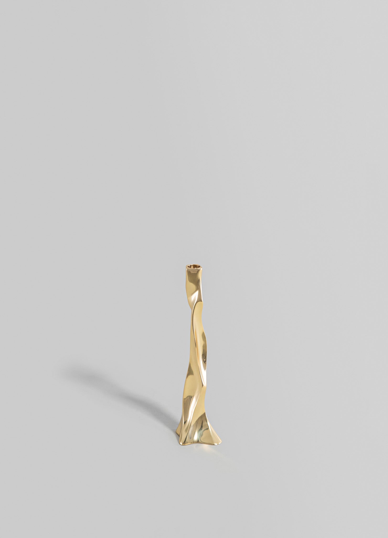 A sculptural gold-colored metal object with an abstract, twisted form, standing upright against a plain light gray background. The piece features a polished, reflective surface with organic, flowing lines.