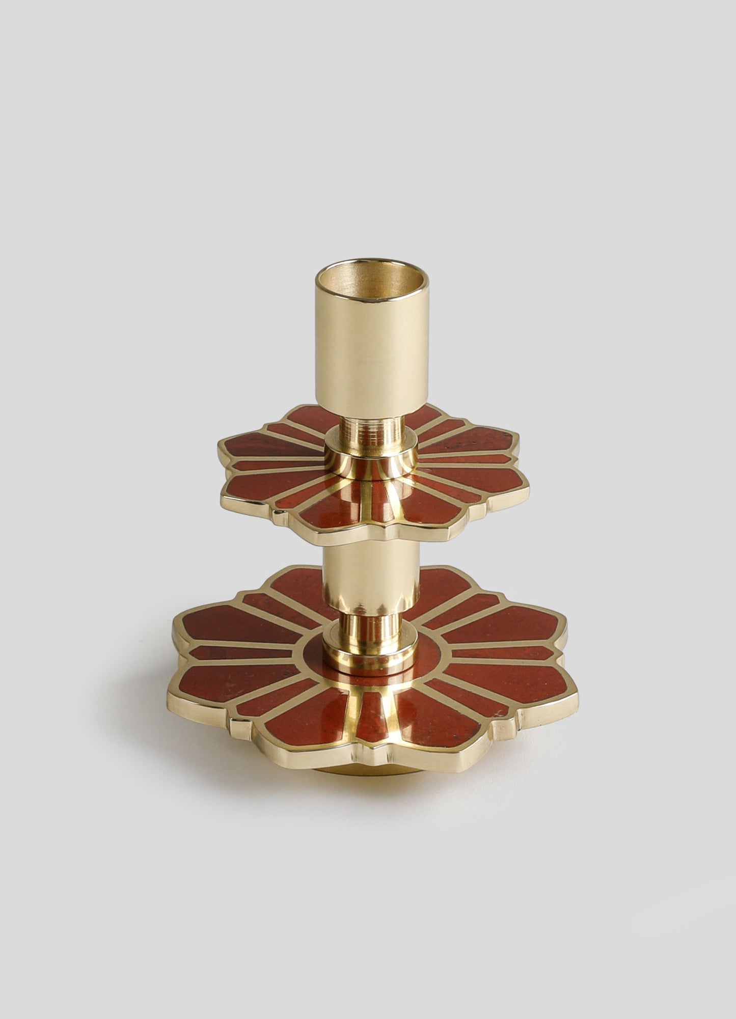 A brass candle holder with a floral motif, featuring layered, petal-shaped bases in red and gold tones, and a cylindrical holder for candles. The design combines contemporary and decorative elements with a reflective metallic finish.