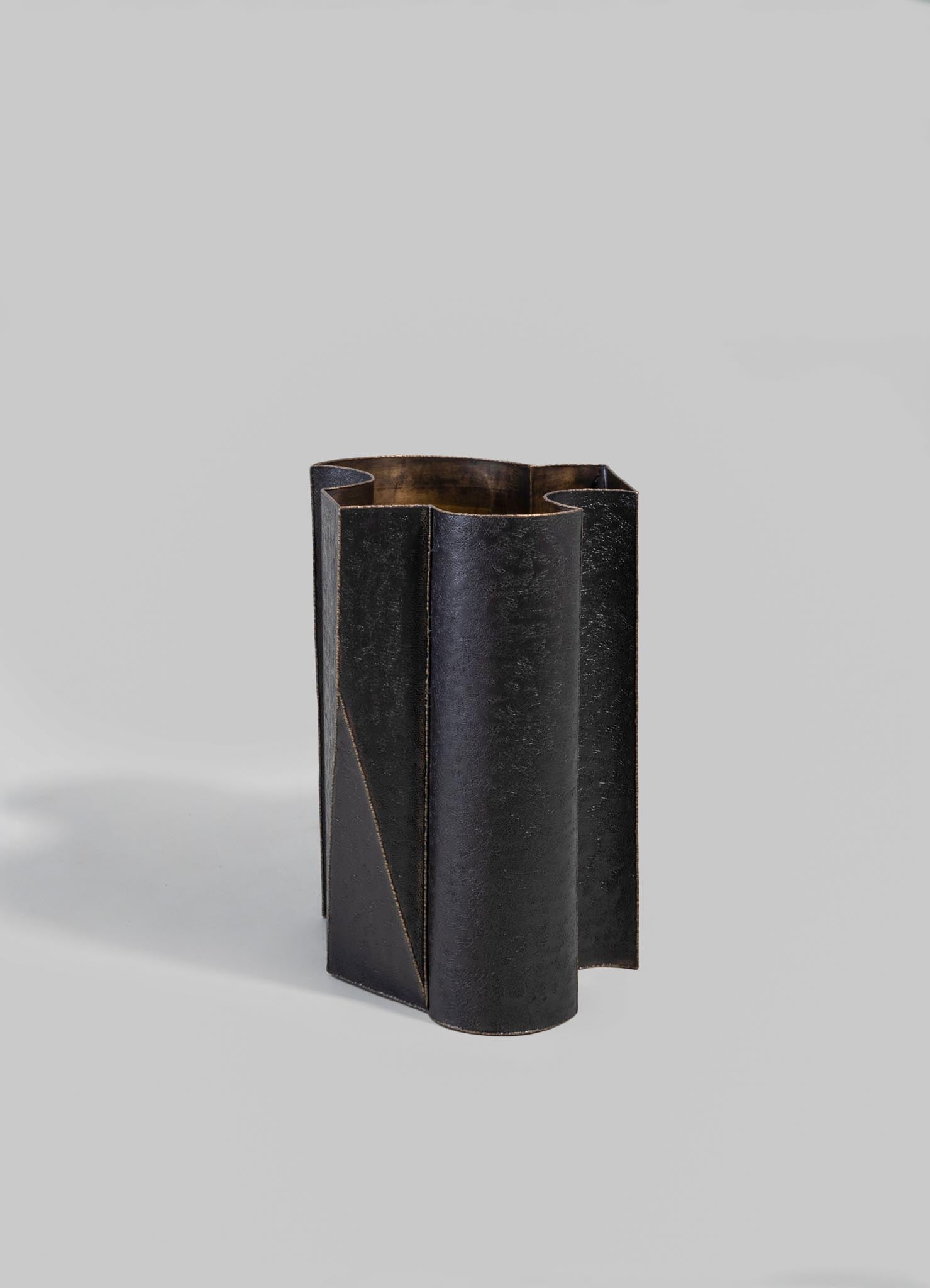 A tall, cylindrical vase made of dark metal with a textured finish, featuring angular cutouts and geometric lines for a modern aesthetic.