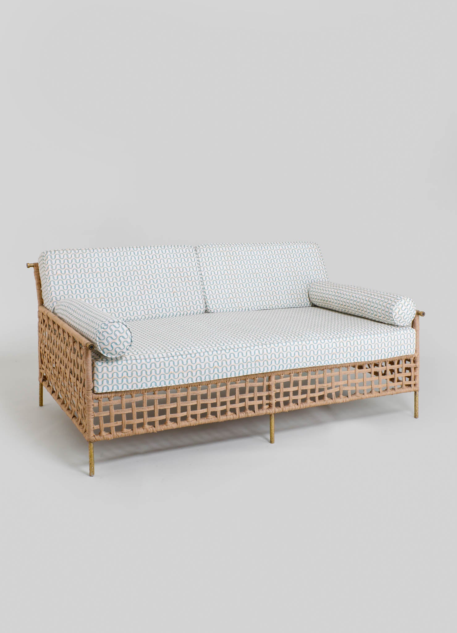 Woven rattan sofa with a light beige finish and upholstered cushions featuring a blue wave pattern, set against a neutral background. The sofa has cylindrical bolster pillows and a contemporary style with natural materials.