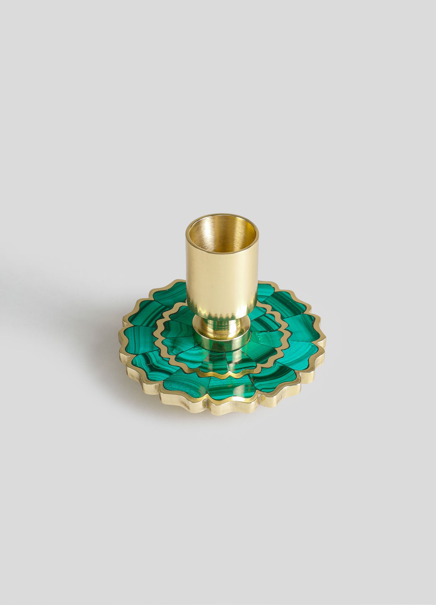A brass candle holder with a cylindrical shape, placed on an intricate green and gold decorative tray with a scalloped edge and floral motif.