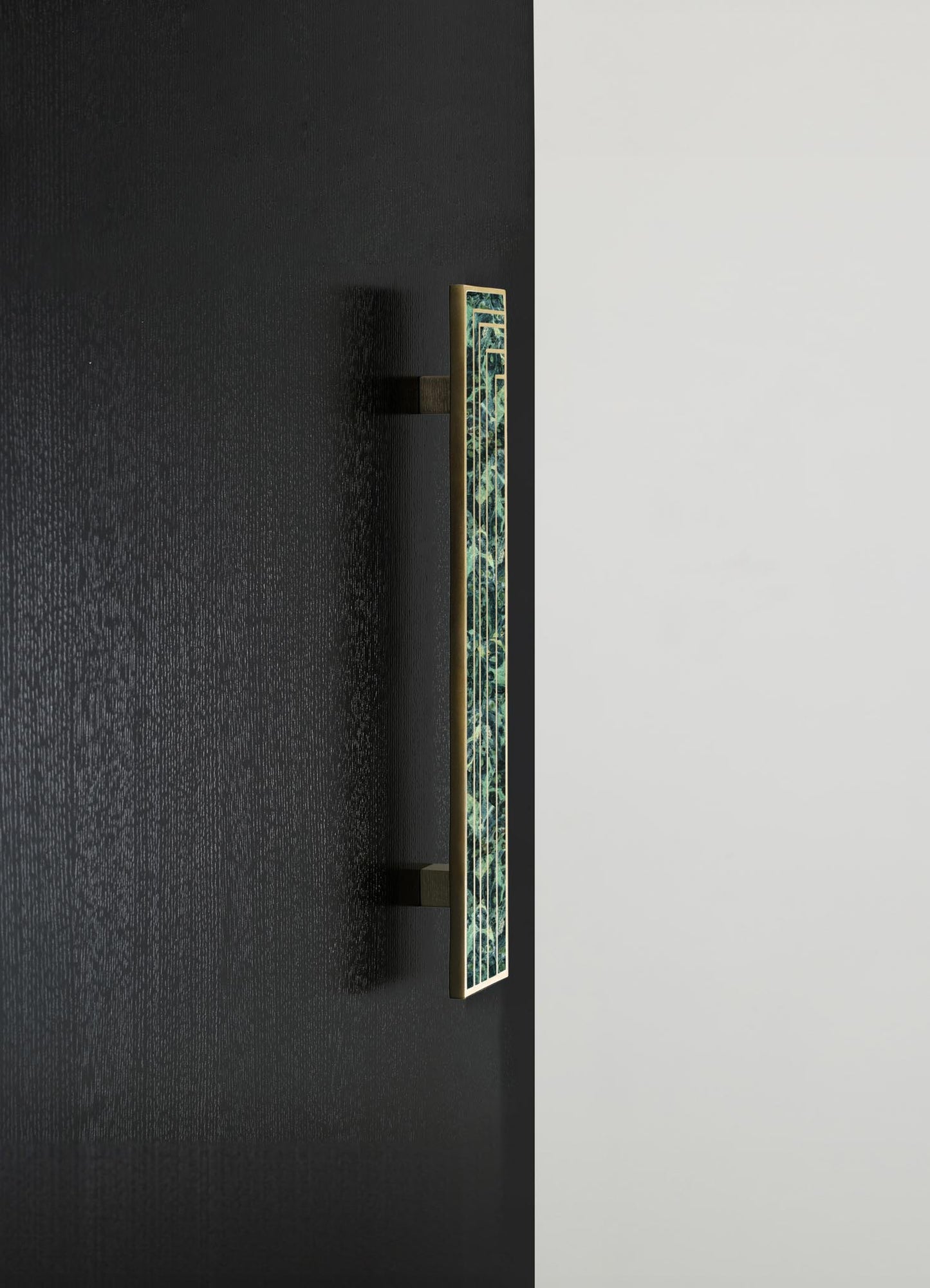 A decorative wall-mounted sconce featuring a narrow rectangular shape with a brass frame and a mosaic pattern of green and black glass, set against a black textured wall and a contrasting white surface.