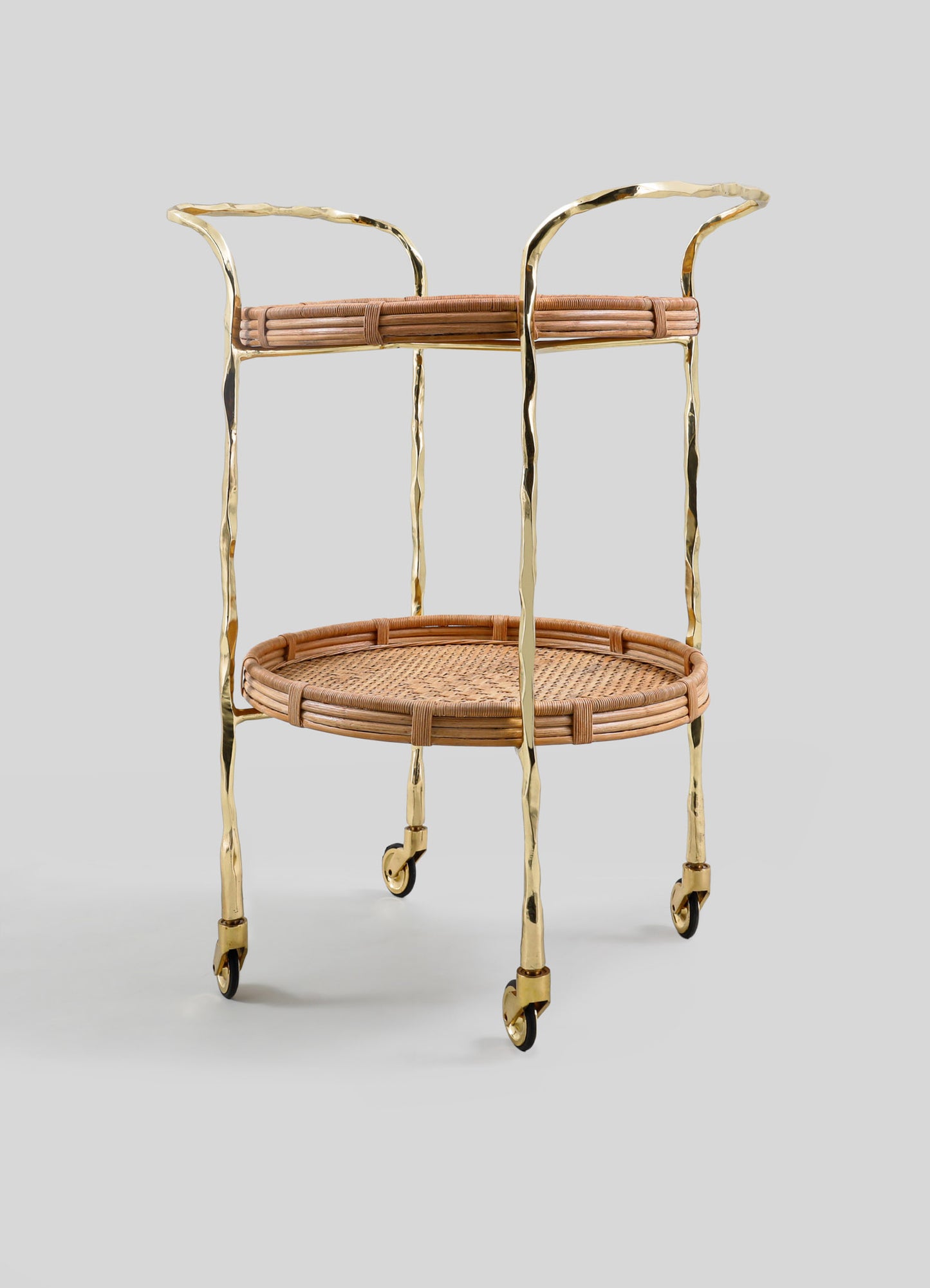The Assam Brass & Cane Trolley by VIYA features a gold metal frame with forged brass rods, curved handles, woven rattan shelves, and four small wheels, shown against a plain light gray background.