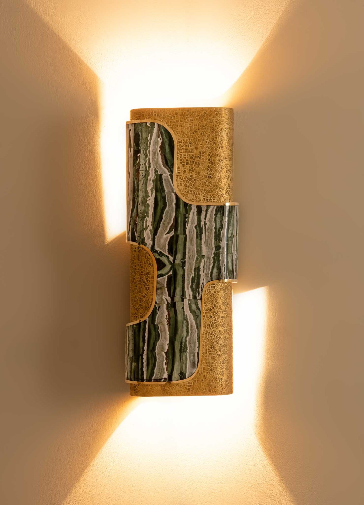 Wall-mounted light fixture with a combination of green and white marble and gold-toned metal accents, emitting warm light and creating a geometric shadow pattern on the wall.