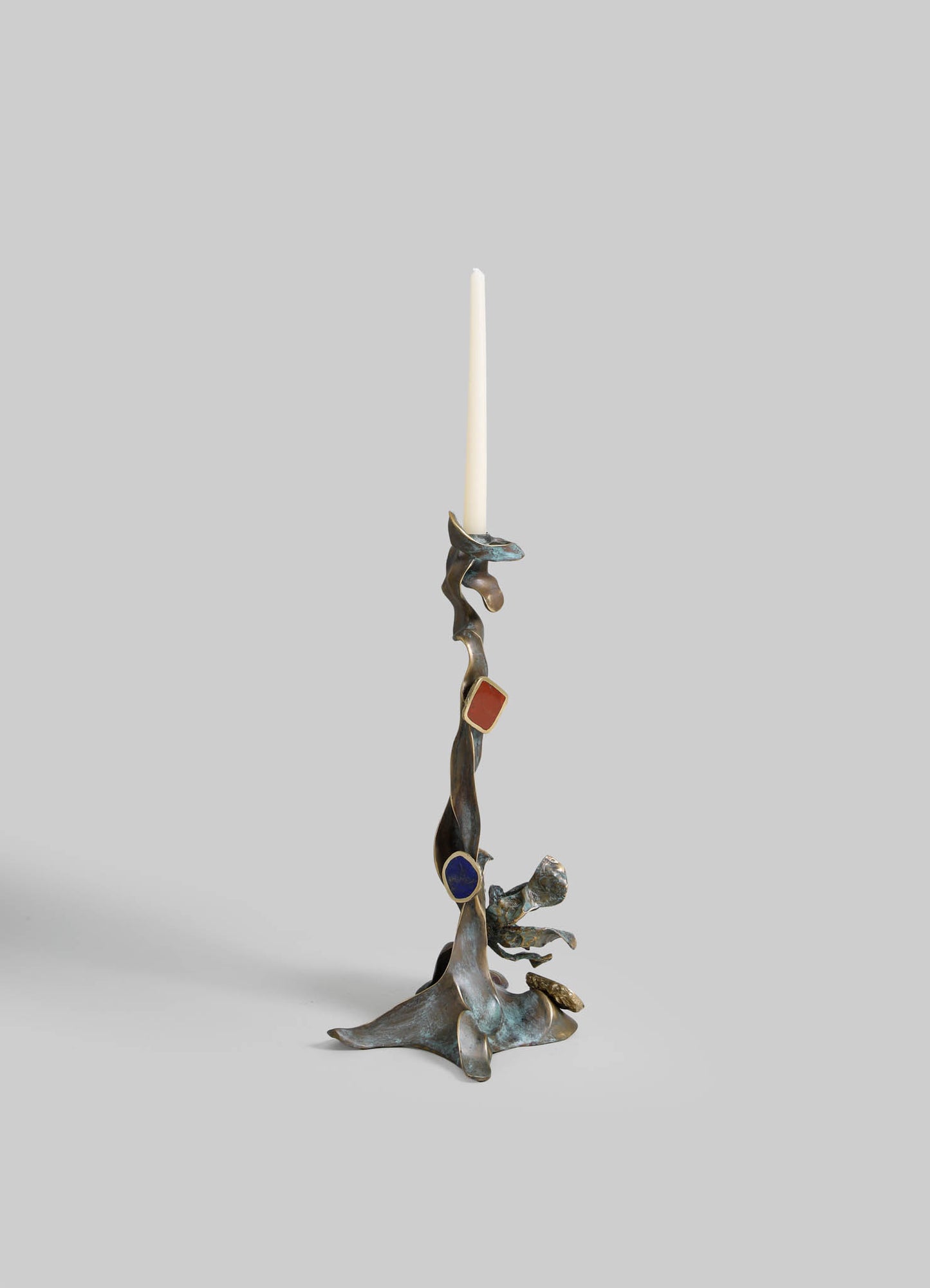 A sculptural candle holder made of metal with an abstract, organic form and a weathered finish, supporting a tall white taper candle. The design features flowing, irregular shapes with hints of blue, red, and gold accents.