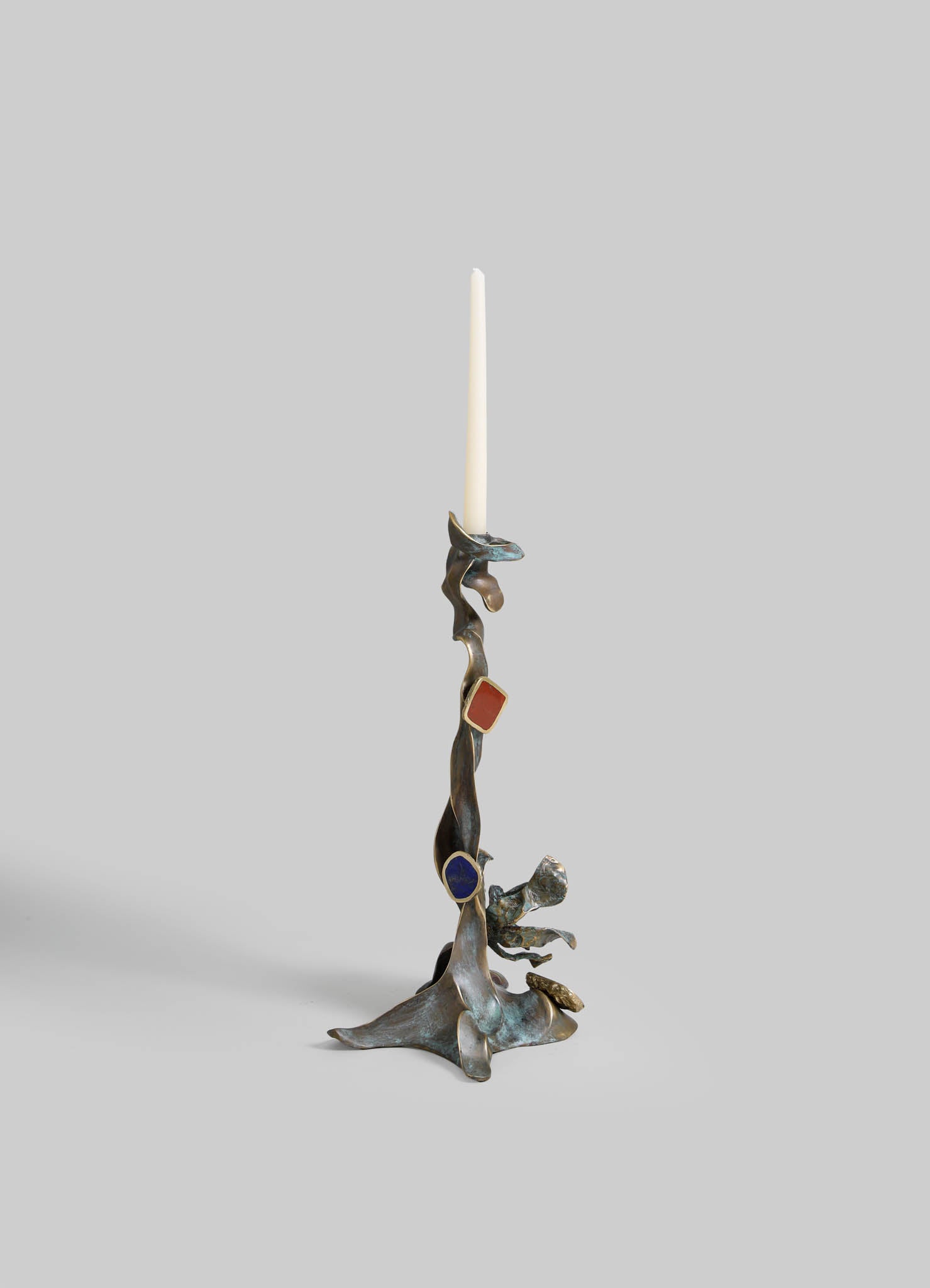 A sculptural candle holder made of metal with an abstract, organic form and a weathered finish, supporting a tall white taper candle. The design features flowing, irregular shapes with hints of blue, red, and gold accents.