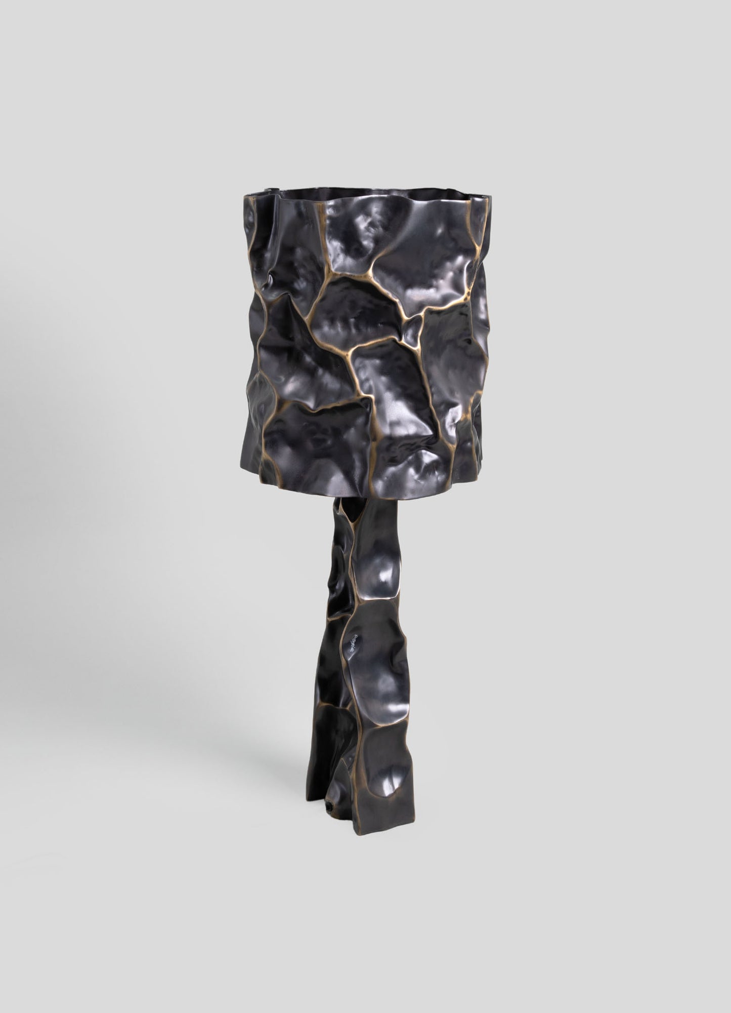 A sculptural table lamp with a textured, irregular surface in a glossy black finish with gold accents, featuring a cylindrical lampshade and a twisted, organic base.