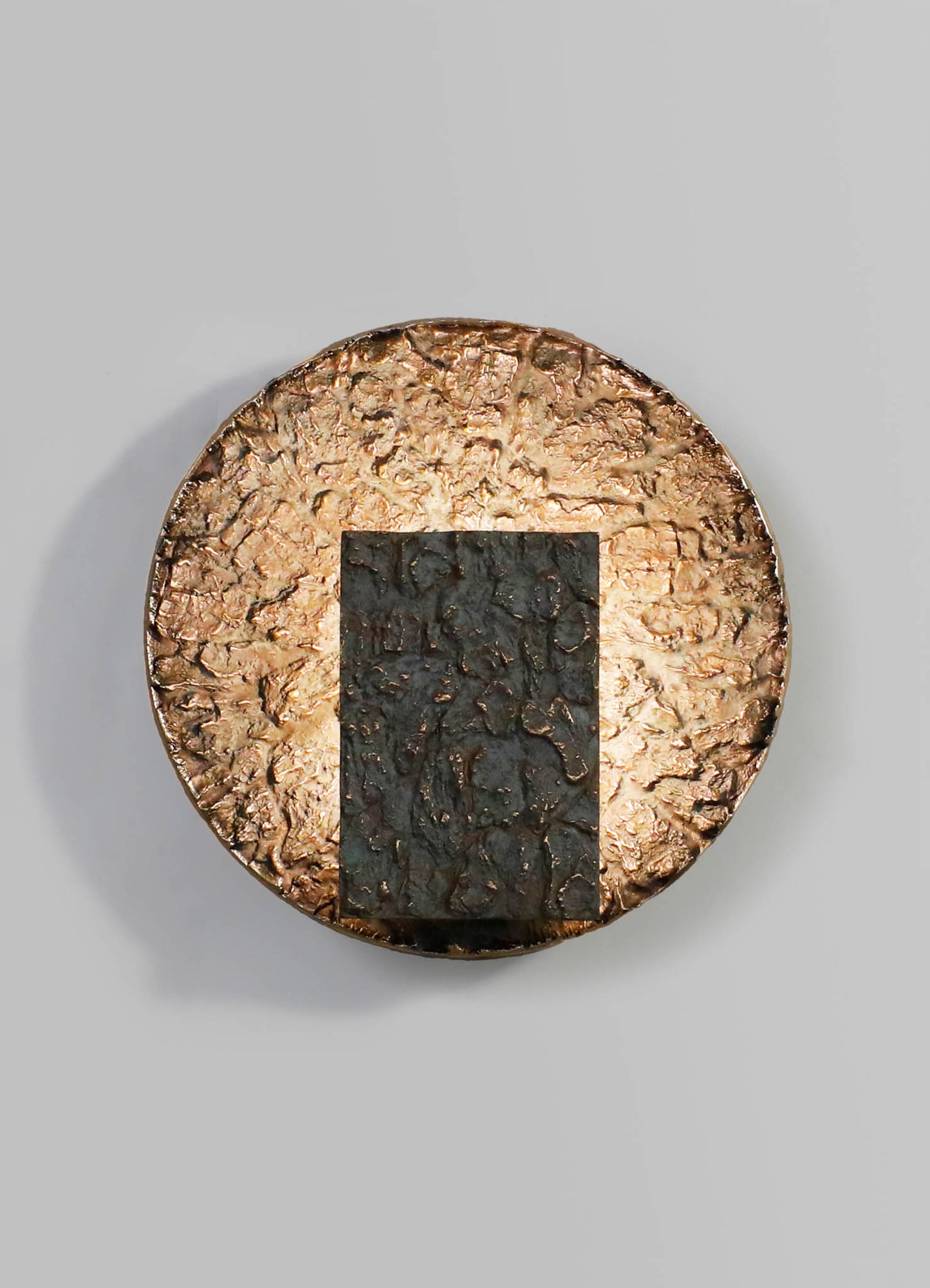 A round decorative object with a textured metallic surface in bronze tones, featuring a rectangular black element with a cracked or distressed finish in the center, set against a plain background.