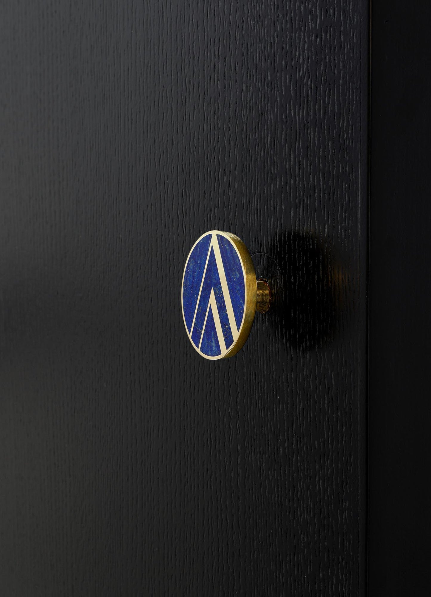 A circular badge or pin with a blue enamel surface and a gold geometric pattern, attached to a textured black surface, likely a piece of furniture or casing. The design features clean, angular lines in a modern style.