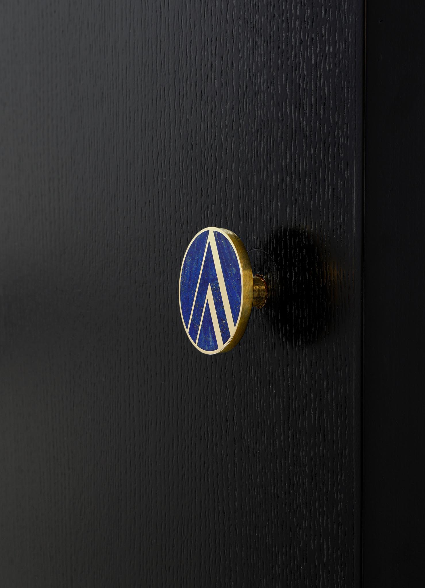 A circular badge or pin with a blue enamel surface and a gold geometric pattern, attached to a textured black surface, likely a piece of furniture or casing. The design features clean, angular lines in a modern style.