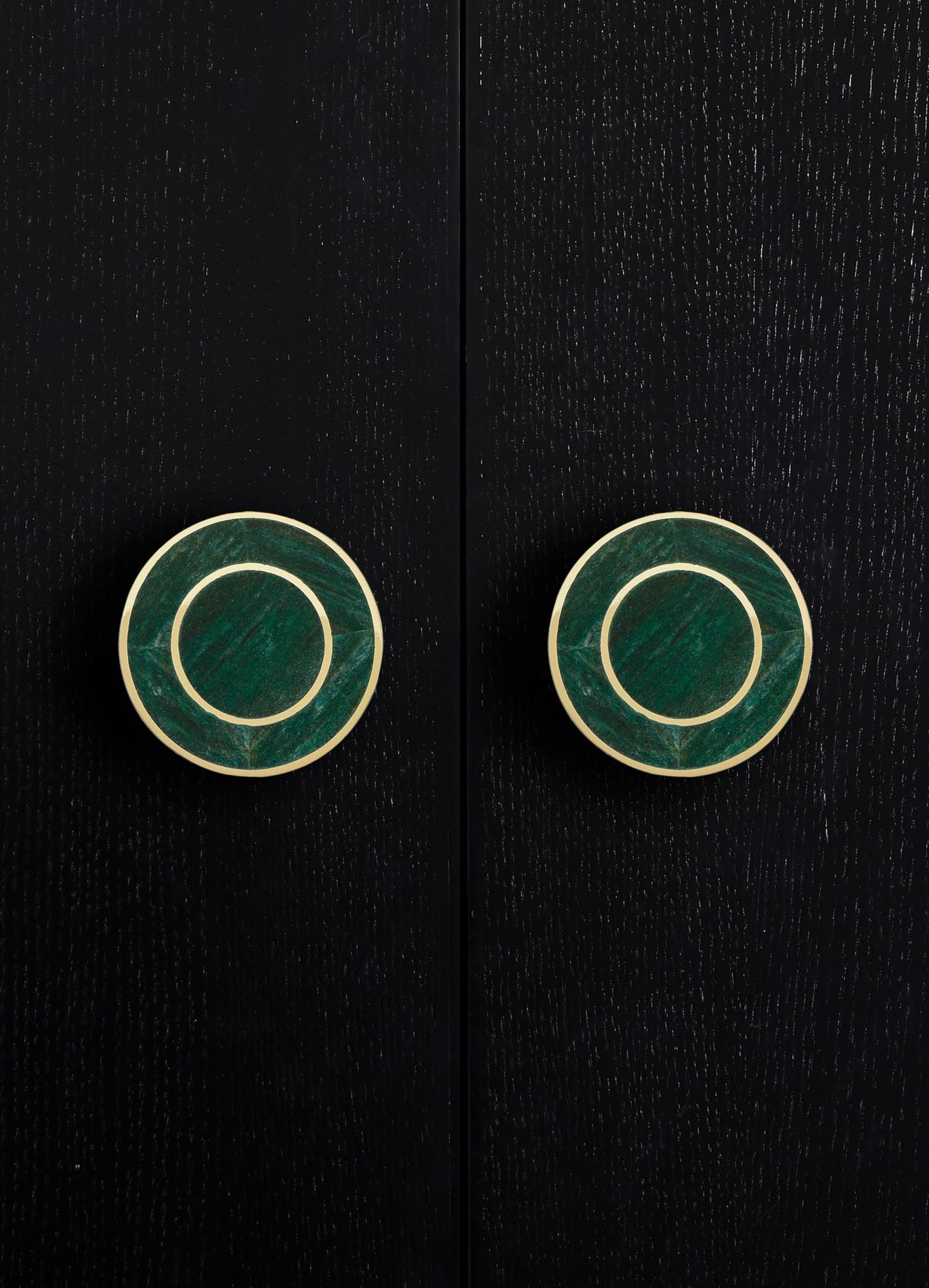 Circular cabinet or drawer handles with green marble or stone inlays and brass or gold-toned borders, mounted on a dark wood surface with a textured grain. The style appears modern and elegant.