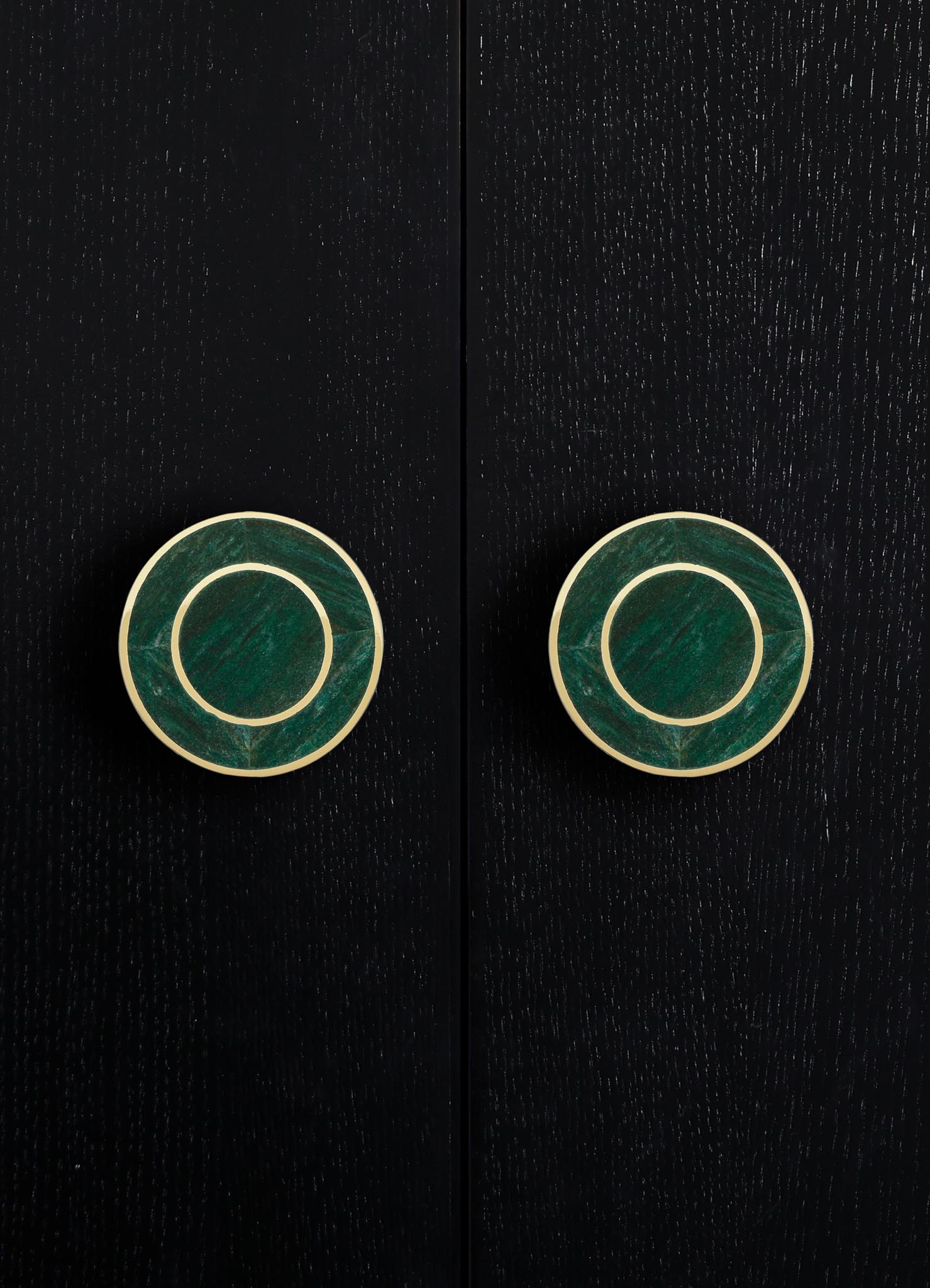 Circular cabinet or drawer handles with green marble or stone inlays and brass or gold-toned borders, mounted on a dark wood surface with a textured grain. The style appears modern and elegant.