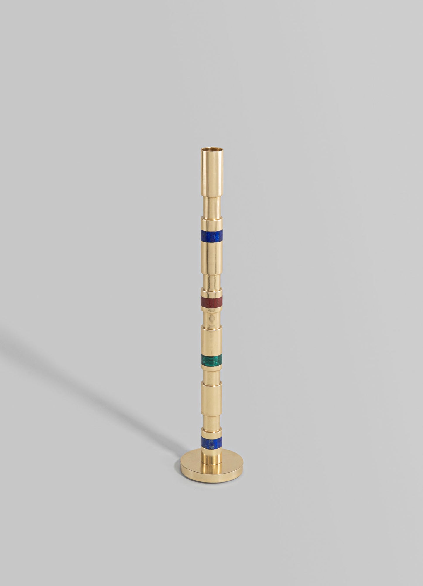 A decorative candlestick holder with a cylindrical brass construction, featuring colorful glass inlays in blue, red, and green, set against a plain gray background.