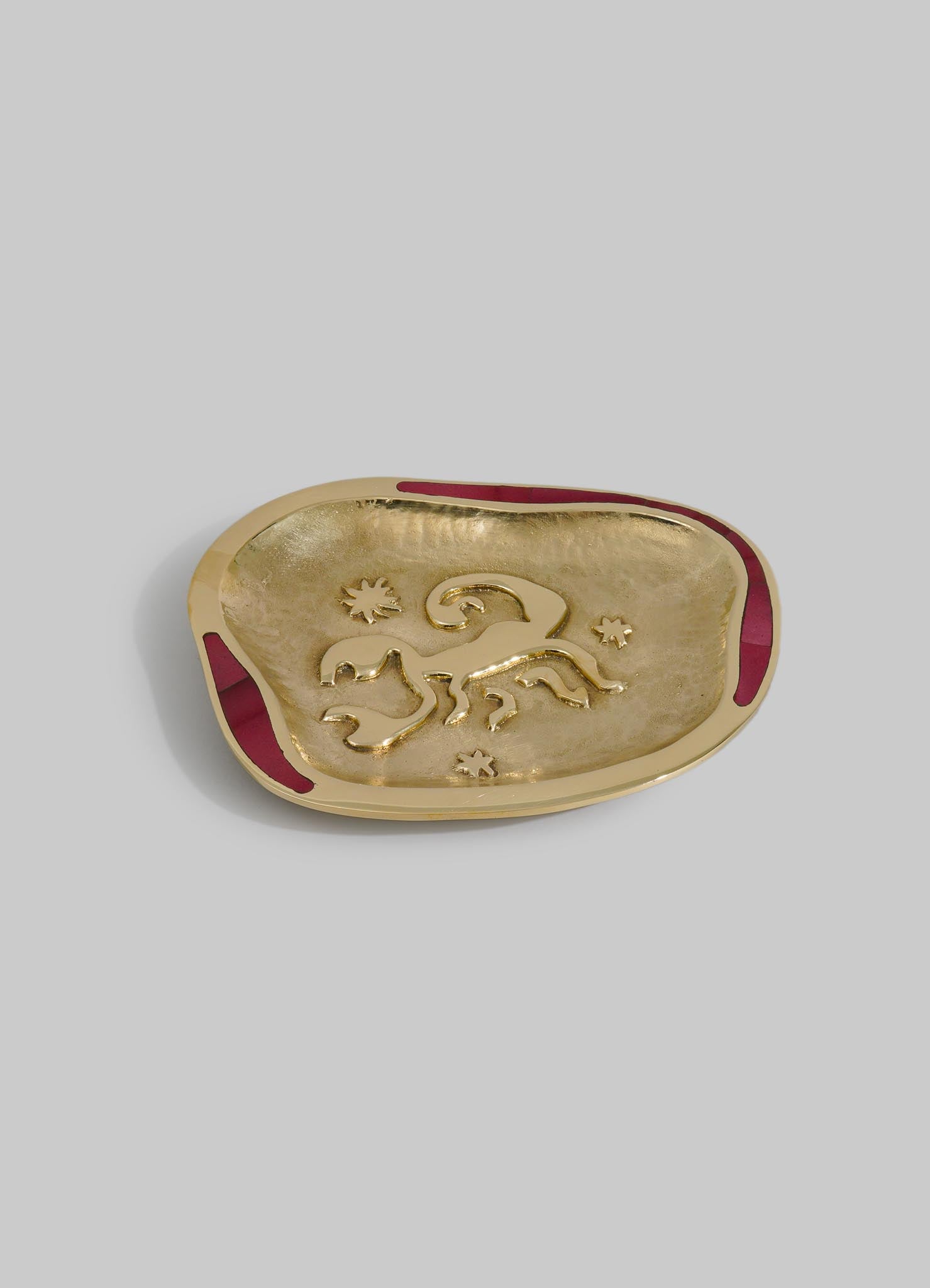 A decorative dish with a gold-toned interior featuring embossed animal motifs and small star accents, bordered with a contrasting red trim. The dish has an irregular oval shape and appears to be made of metal with a sleek, polished finish.