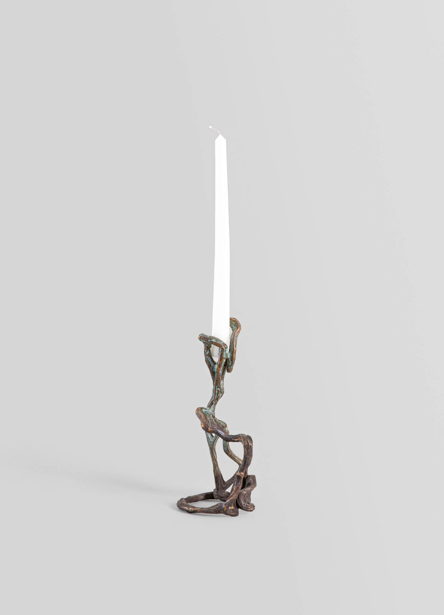A candlestick holder made of metal with an organic, twisted design resembling intertwined branches or roots, featuring a dark, weathered finish. It holds a tall, white taper candle and exhibits a rustic, handcrafted style.