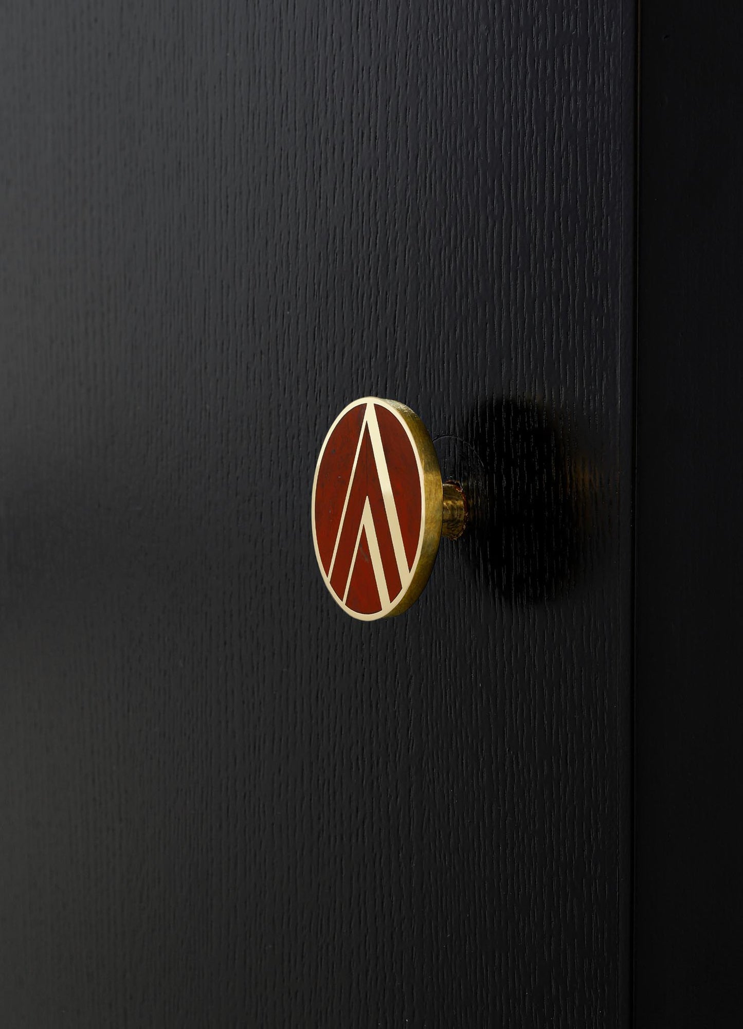 A close-up of a circular decorative knob with a geometric pattern in white and red, mounted on a black textured wooden surface. The knob has a metallic gold edge, adding a subtle contrast to the matte black background.