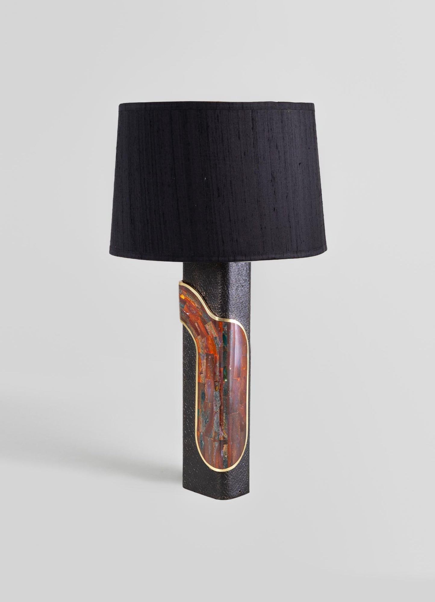 A table lamp with a black fabric shade and a textured black base featuring an colorful, abstract inlay with warm red, orange, and green tones, and gold trim.