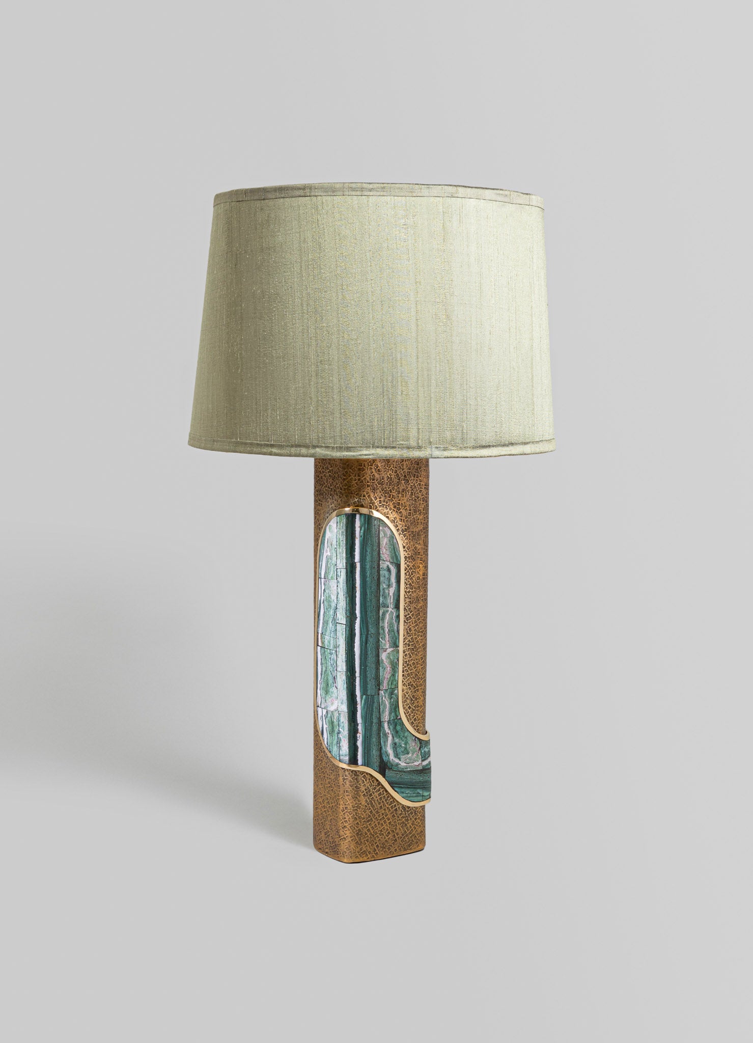 A table lamp with a cylindrical base featuring textured metallic gold finish and a green marble inlay, topped with a beige fabric lampshade. The style is contemporary with a focus on luxury and mixed materials.