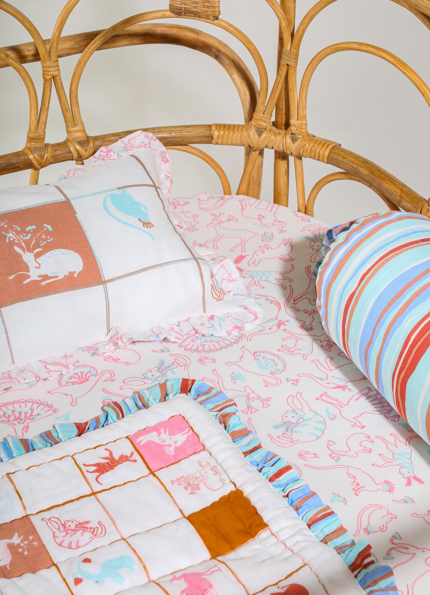 A bed with a rattan headboard featuring curved and woven design elements, dressed in fabric with colorful, playful animal and floral motifs. The bedding includes a patchwork pillow with embroidered animals, a pillow with a striped pattern in shades of orange, blue, and white, and a bedsheet with a pink and light blue animal print.