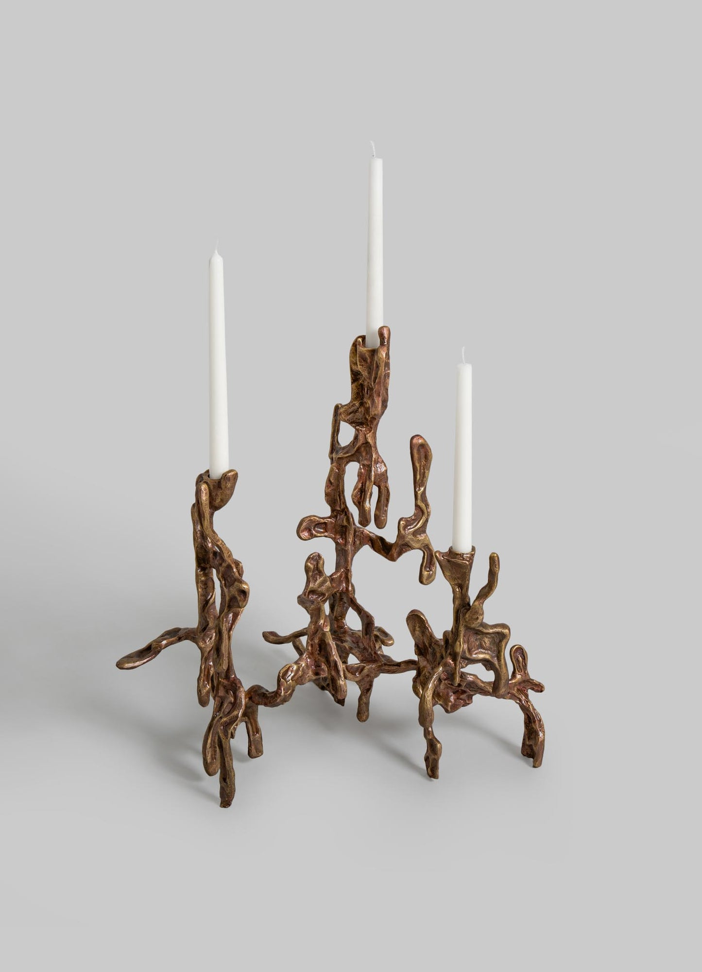 A decorative brass candle holder with an abstract, organic design resembling twisted roots or branches, holding three white taper candles against a neutral background.