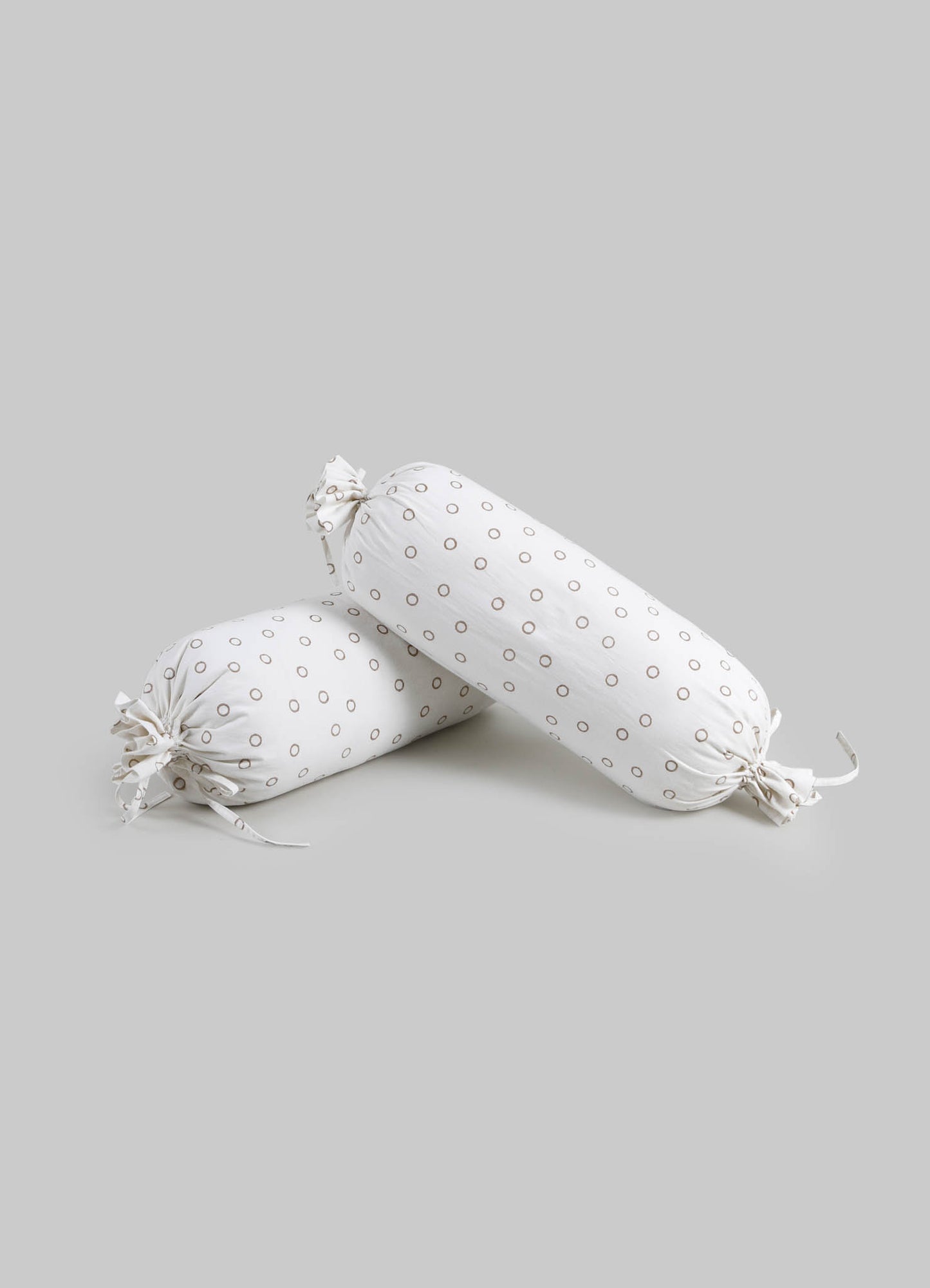 Two cylindrical cushions made of white fabric with small brown circular patterns, tied at each end with matching fabric ties, set against a light gray background.