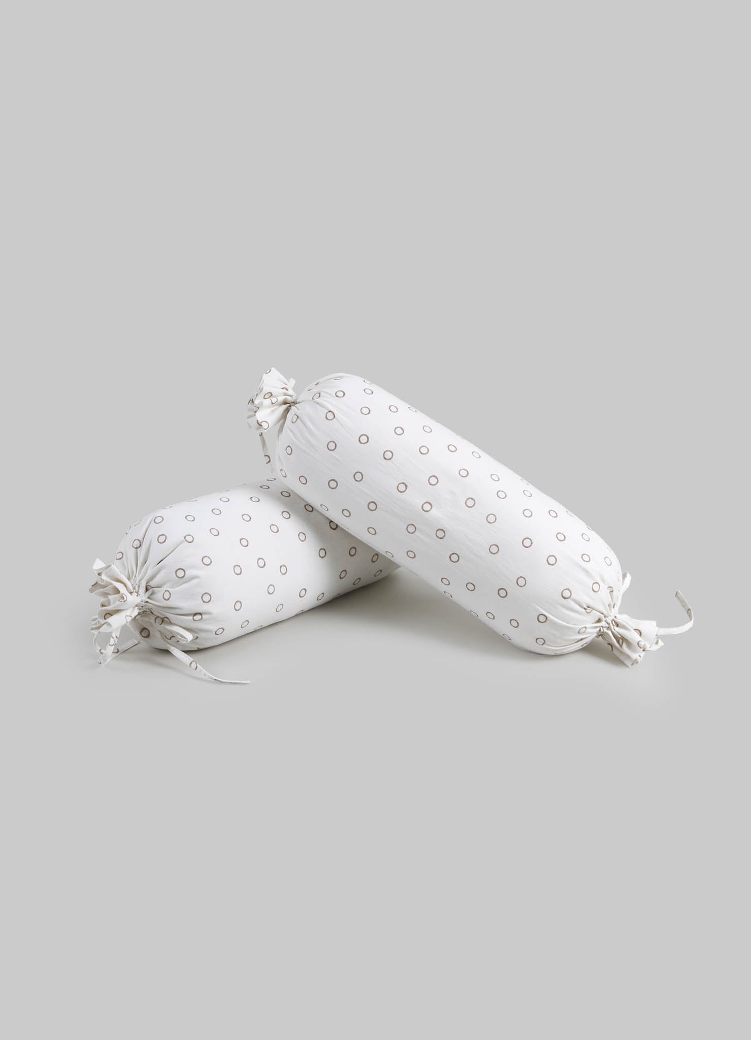 Two cylindrical cushions made of white fabric with small brown circular patterns, tied at each end with matching fabric ties, set against a light gray background.