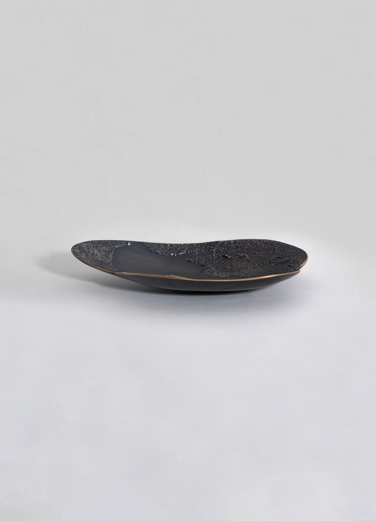 A shallow ceramic dish with an organic, elongated shape, featuring a black glazed interior with textured, speckled patterns and a matte beige exterior. The design has a contemporary and artisanal style.