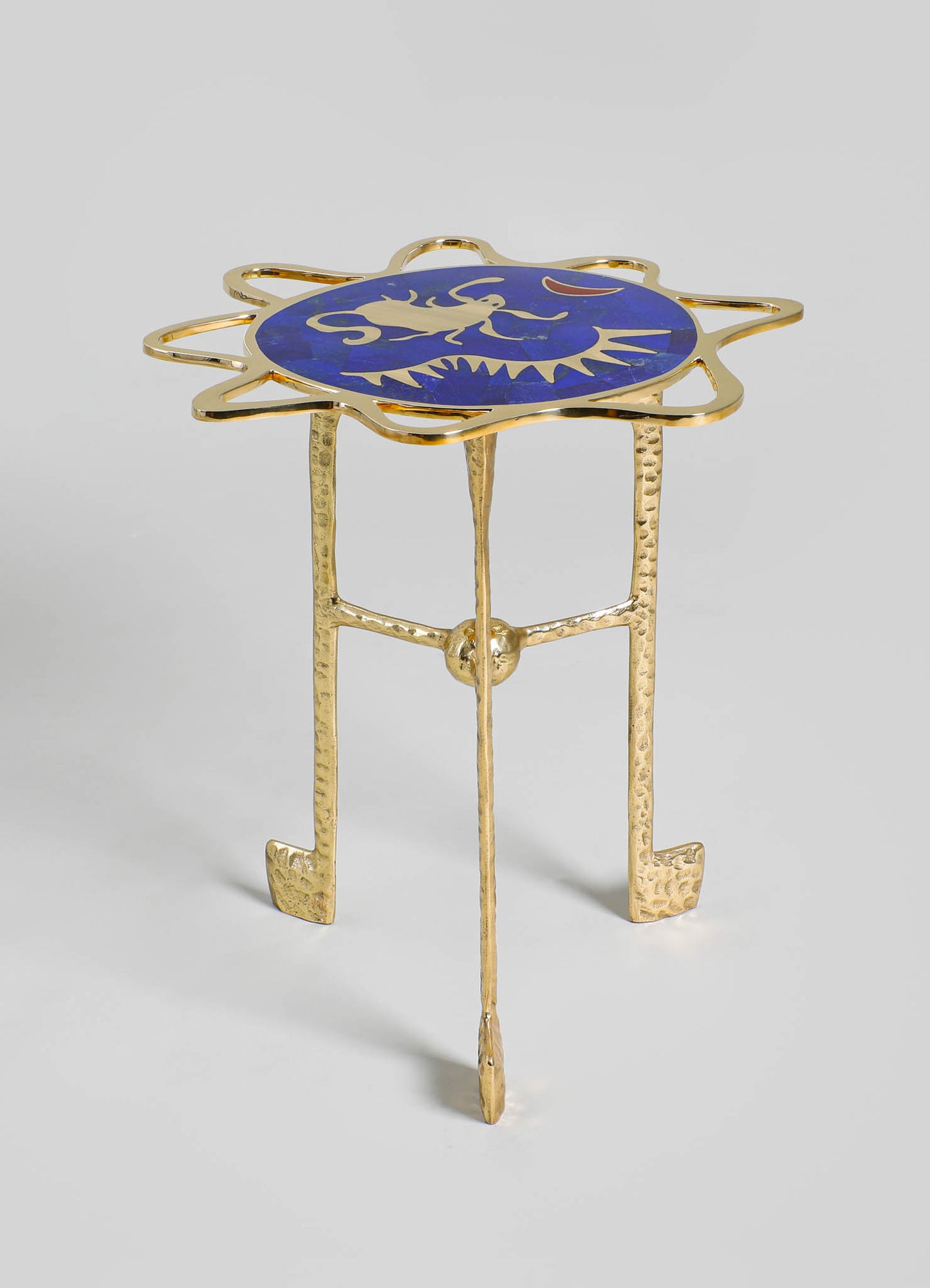 A gold-toned metal side table with a decorative blue ceramic top featuring a dragon motif and a red crescent shape, styled with a handcrafted and contemporary aesthetic. The table has three textured legs with a hammered finish.