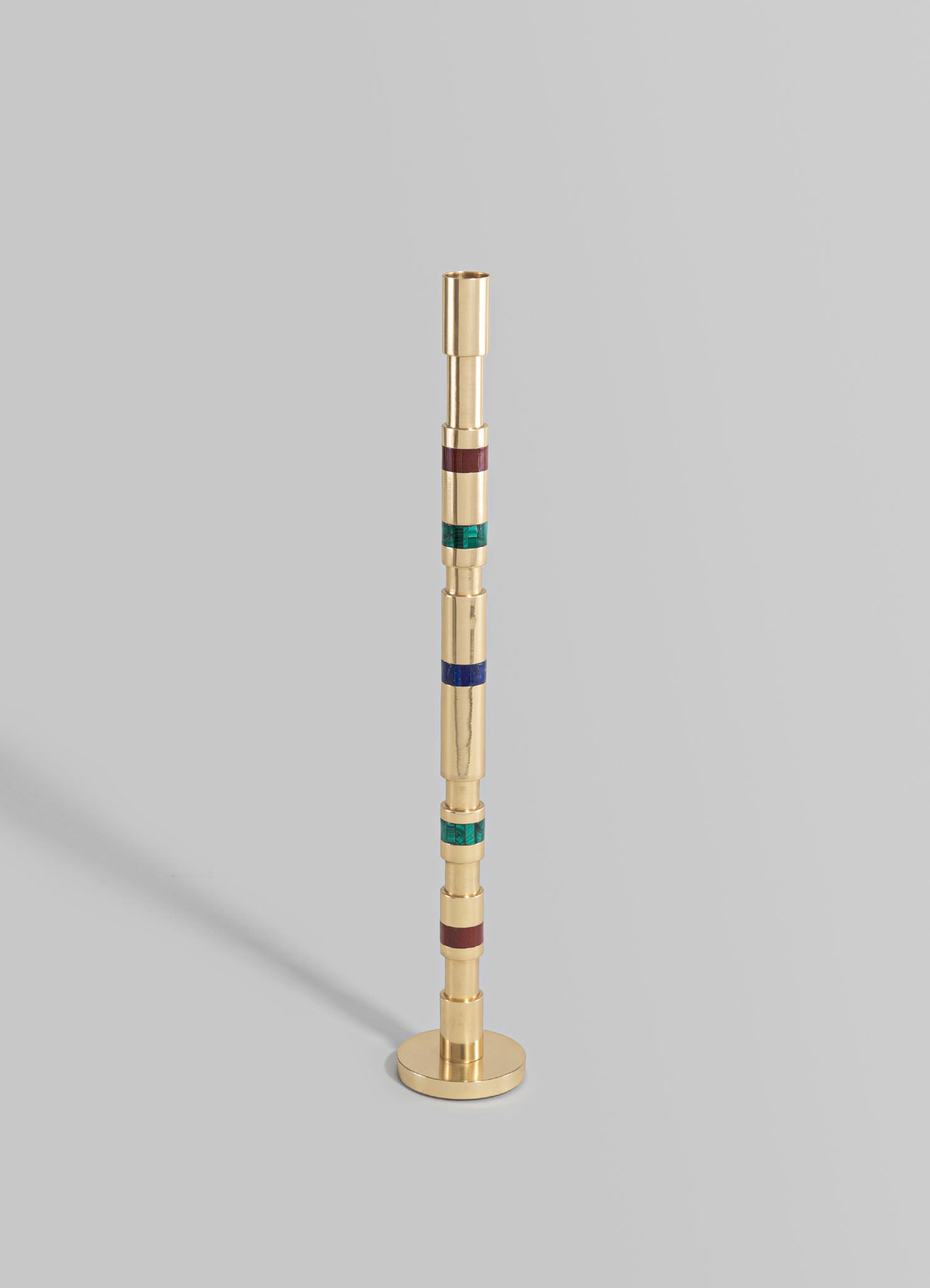 A tall, cylindrical brass sculpture with colorful horizontal bands in burgundy, teal, and blue, featuring a round base and a smooth polished finish.